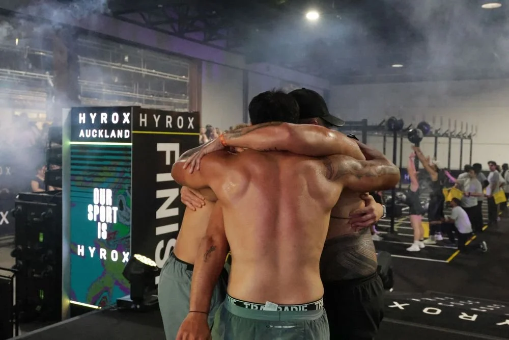 Three shirtless men hugging each other in a gym or CrossFit setting, with other people working out in the background.