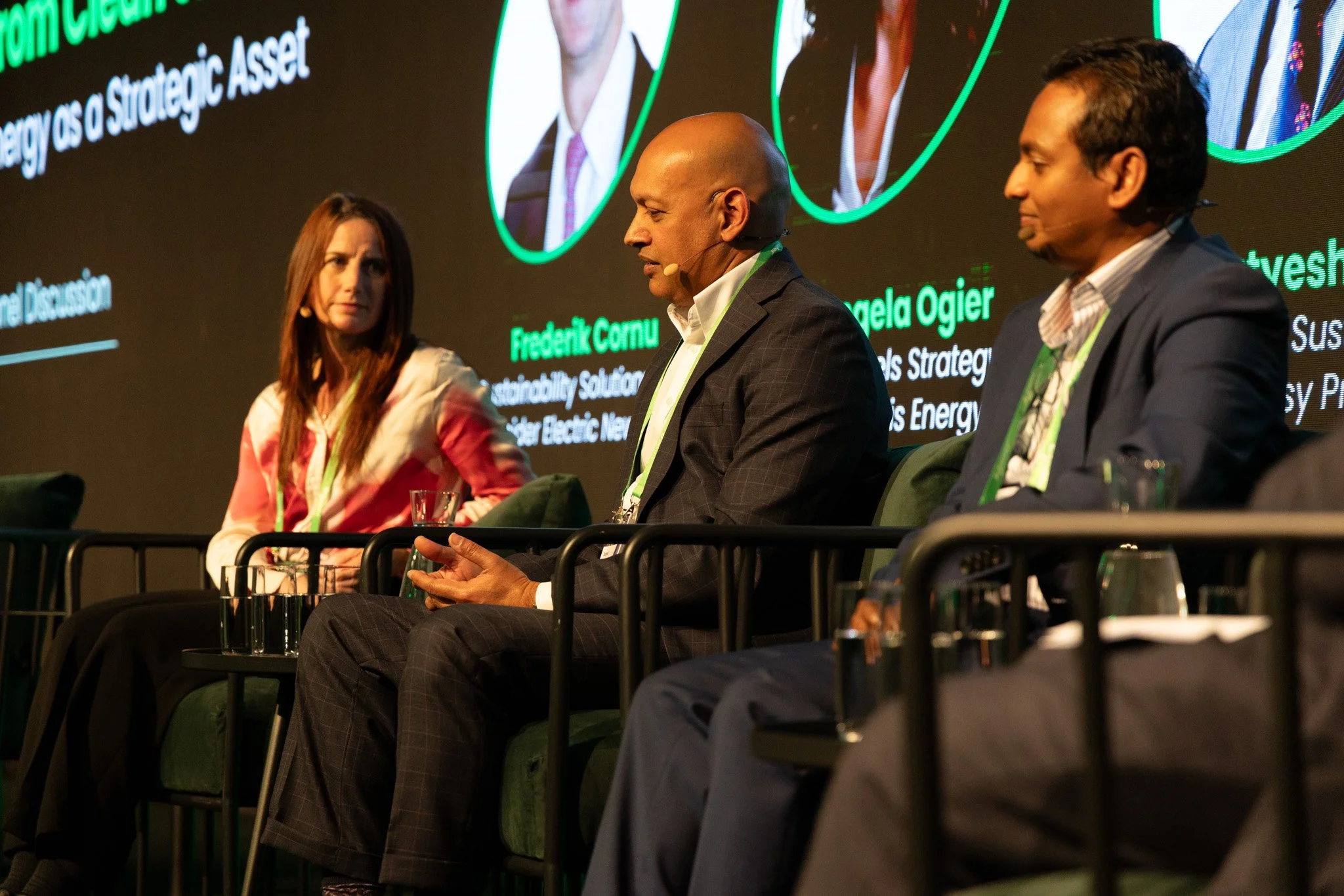 Three people speaking at a panel discussion on stage, with a large screen behind them displaying text and images.