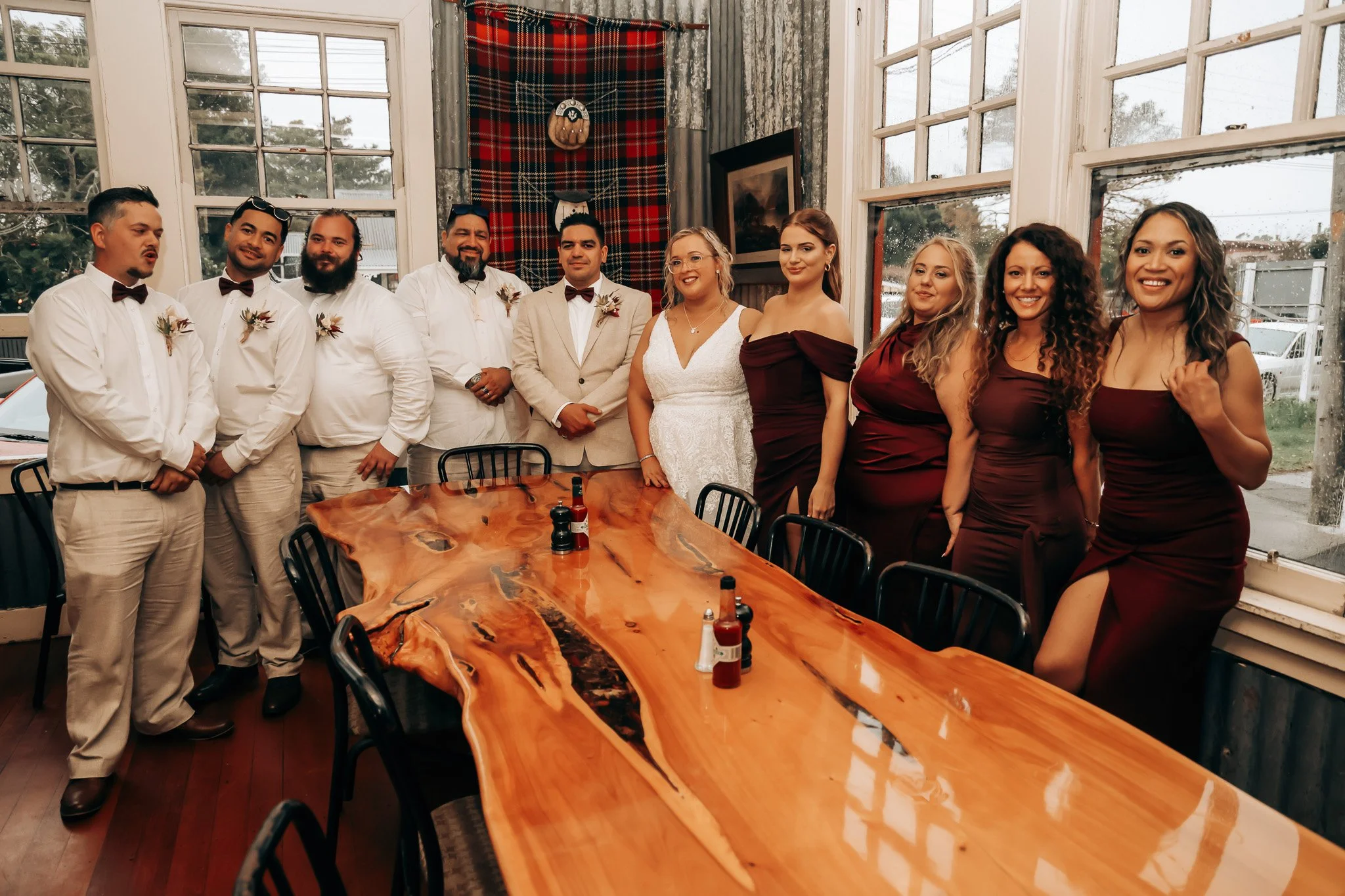 A group of ten people, including men and women, gathered in a rustic indoor setting with large windows, posing for a photo. The group includes both casual and formal attire, and there's a wooden table in the foreground with condiments.