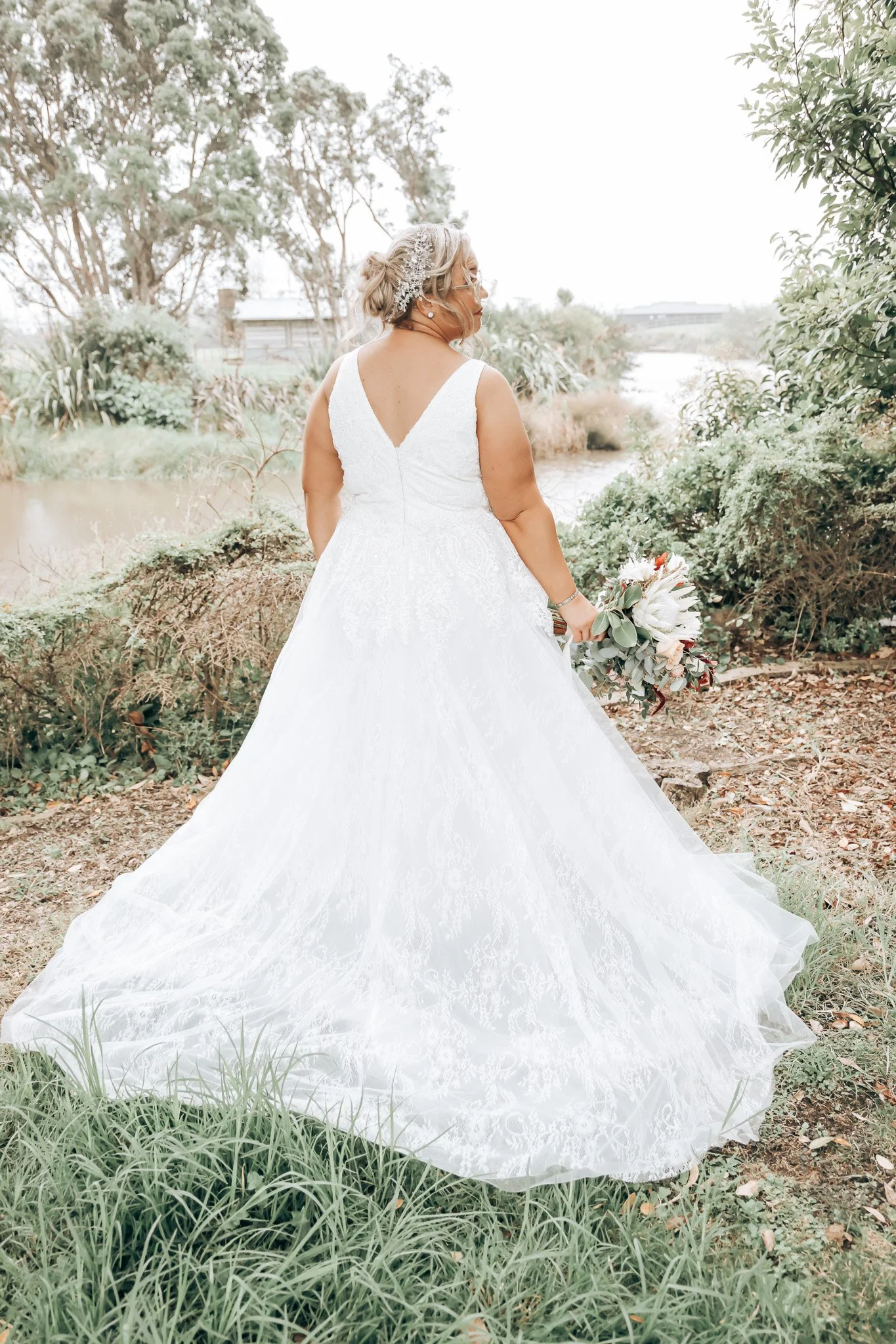 A bride in a white lace wedding gown holding a bouquet, standing outdoors by a pond with trees and shrubbery around.