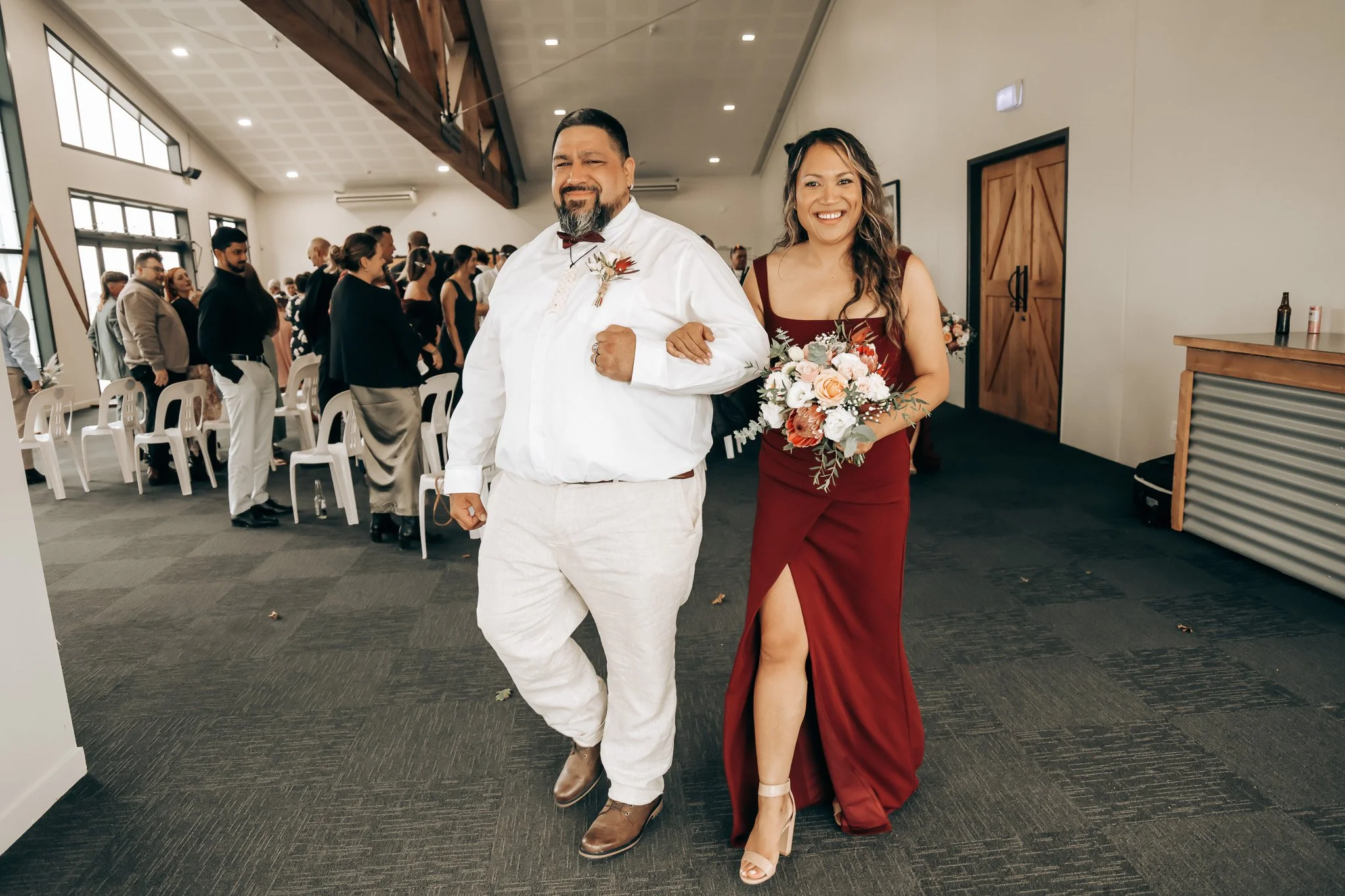 A woman in a burgundy gown holding a bouquet and a man in white attire walking down an aisle at a wedding ceremony, with guests seated and standing in the background.