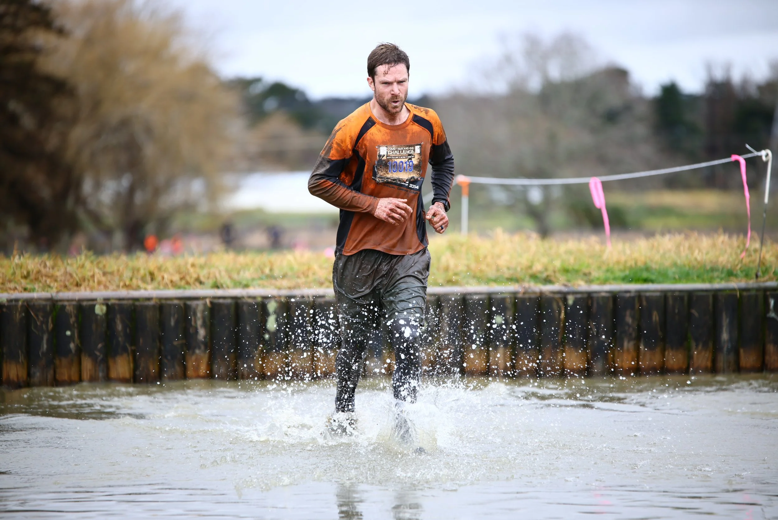 A man running through a muddy water obstacle course during a race, wearing a race bib and dirt-stained clothes.