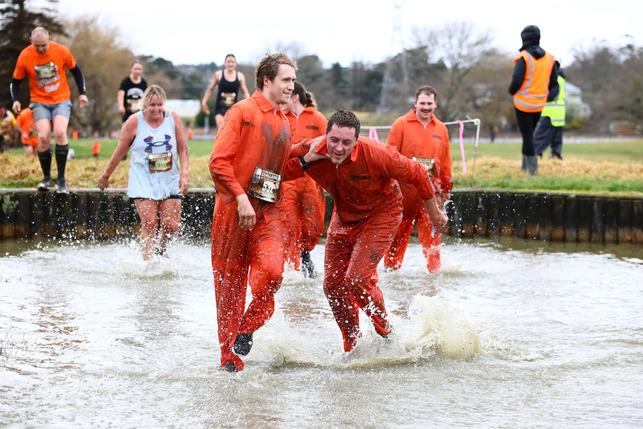 People participating in a muddy outdoor obstacle course race, crossing a water obstacle, wearing athletic gear, some with race bibs, with one person assisting another in the water, and a volunteer in a high-visibility vest overseeing the event.