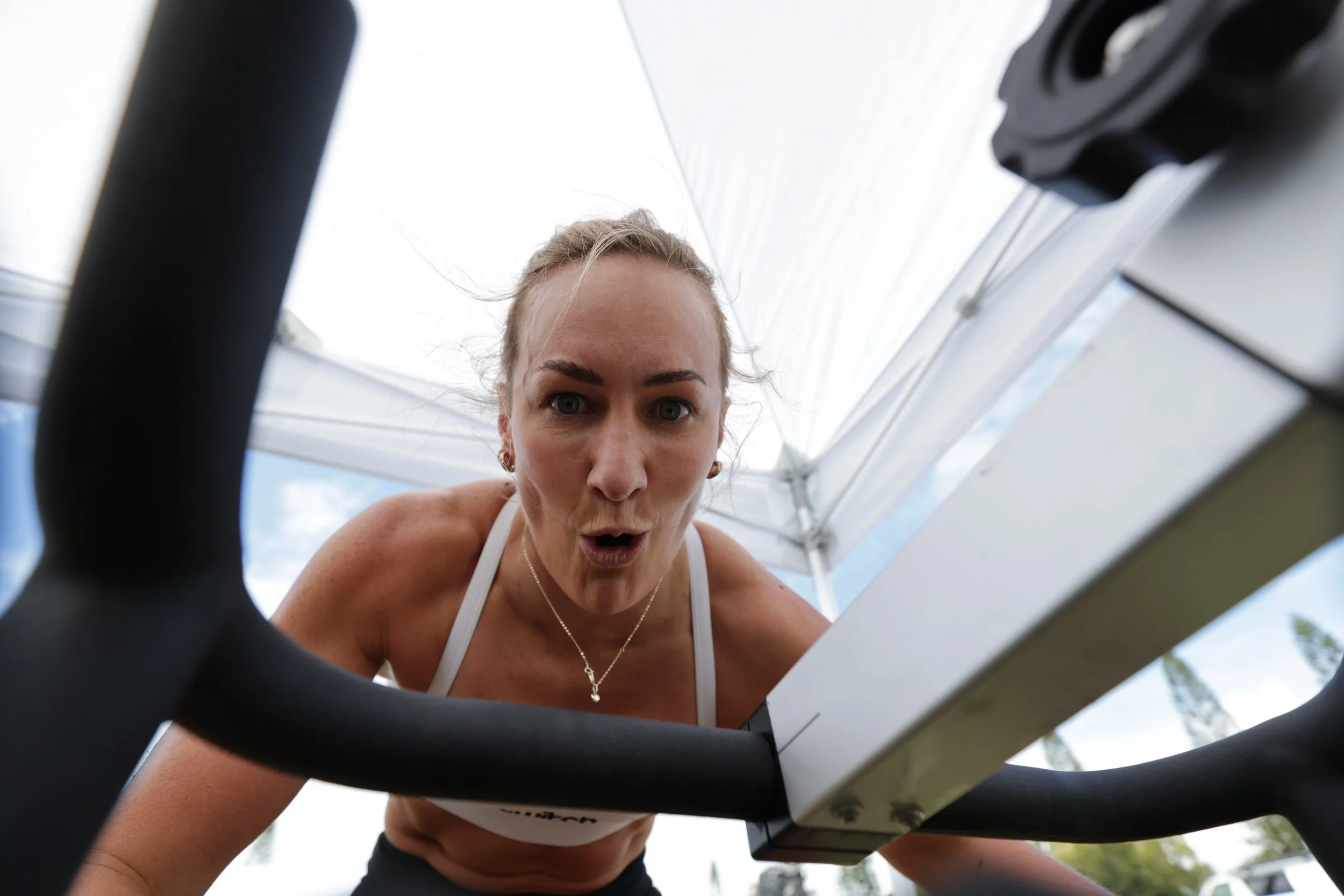 A woman with blonde hair tied back, wearing a white tank top and a necklace, is looking down at a treadmill from below, with a surprised or excited expression.
