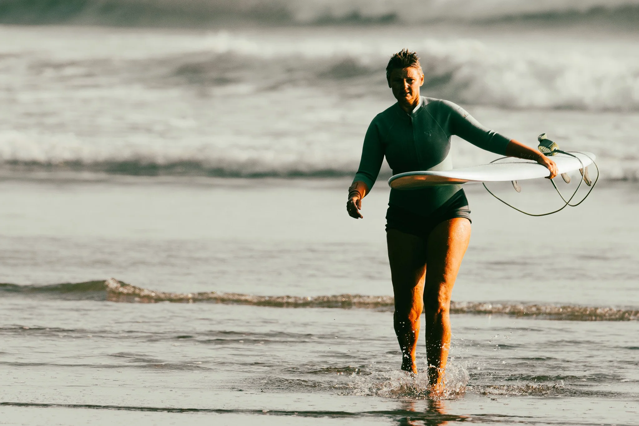 A woman walking out of the ocean, holding a surfboard in her right hand, wearing a wetsuit, with ocean waves in the background.