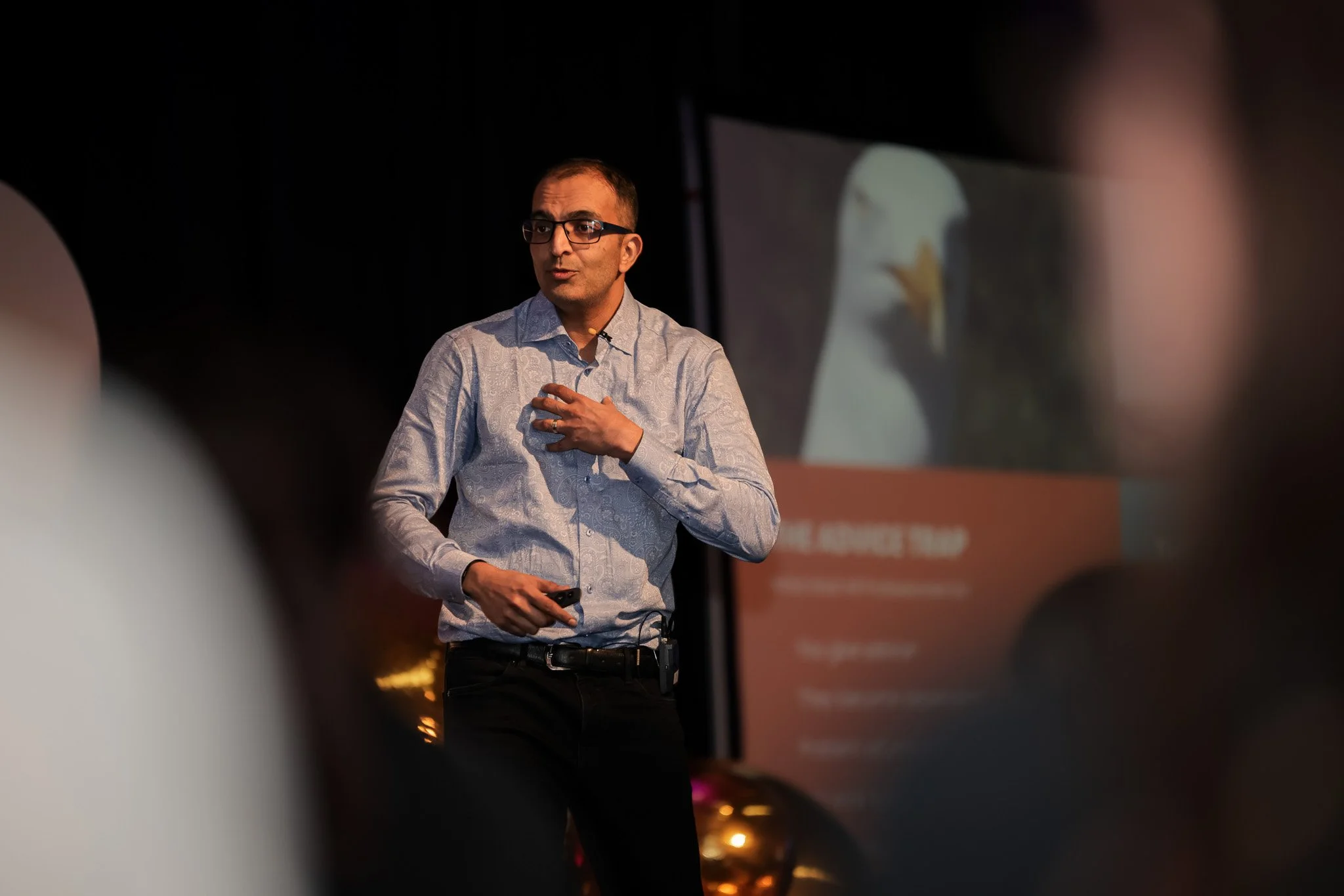 A man with glasses in a patterned shirt giving a presentation or speech, standing in front of a projection screen in a dark room.