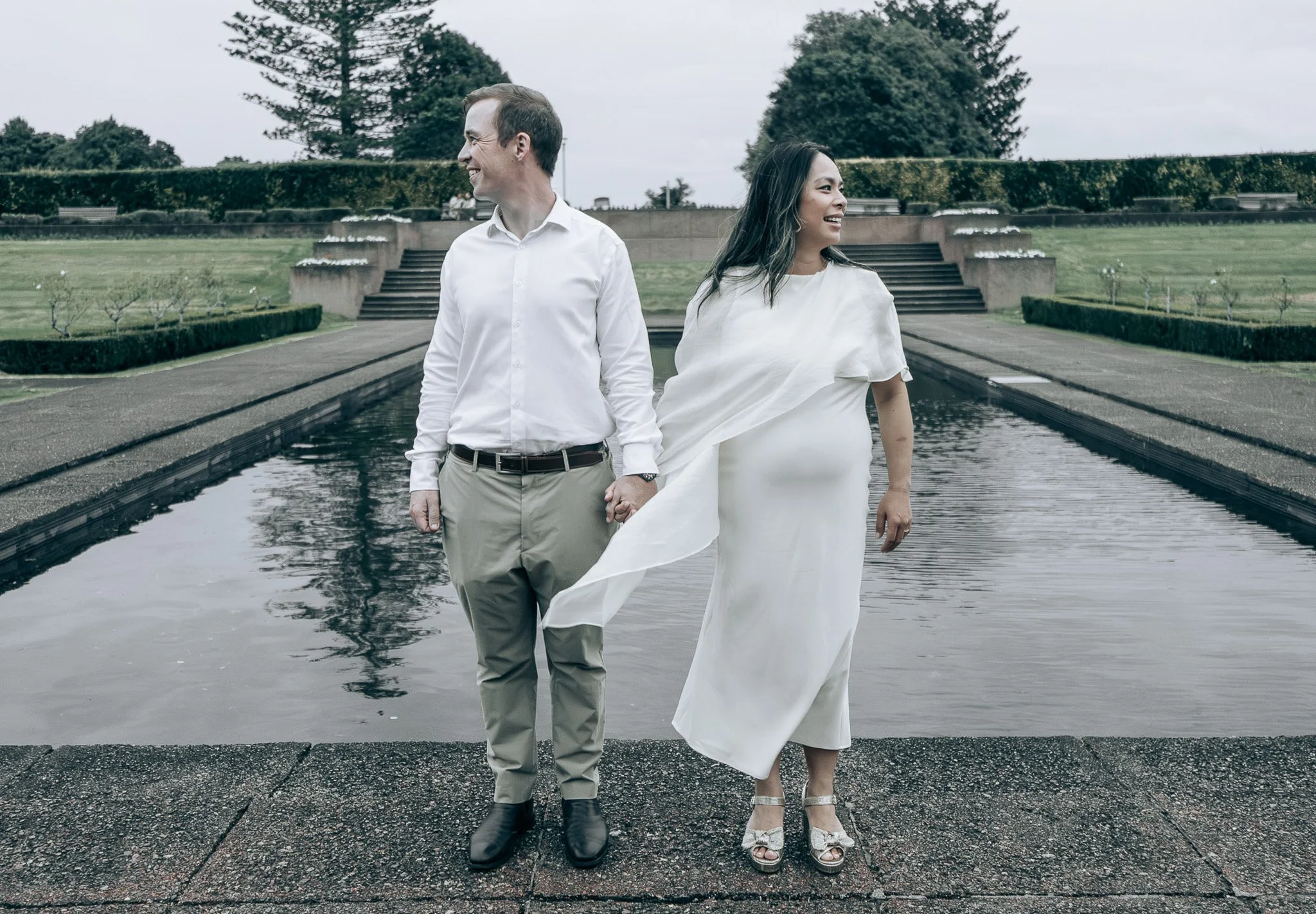 A man and woman holding hands and smiling, standing near a fountain outdoors on a cloudy day.