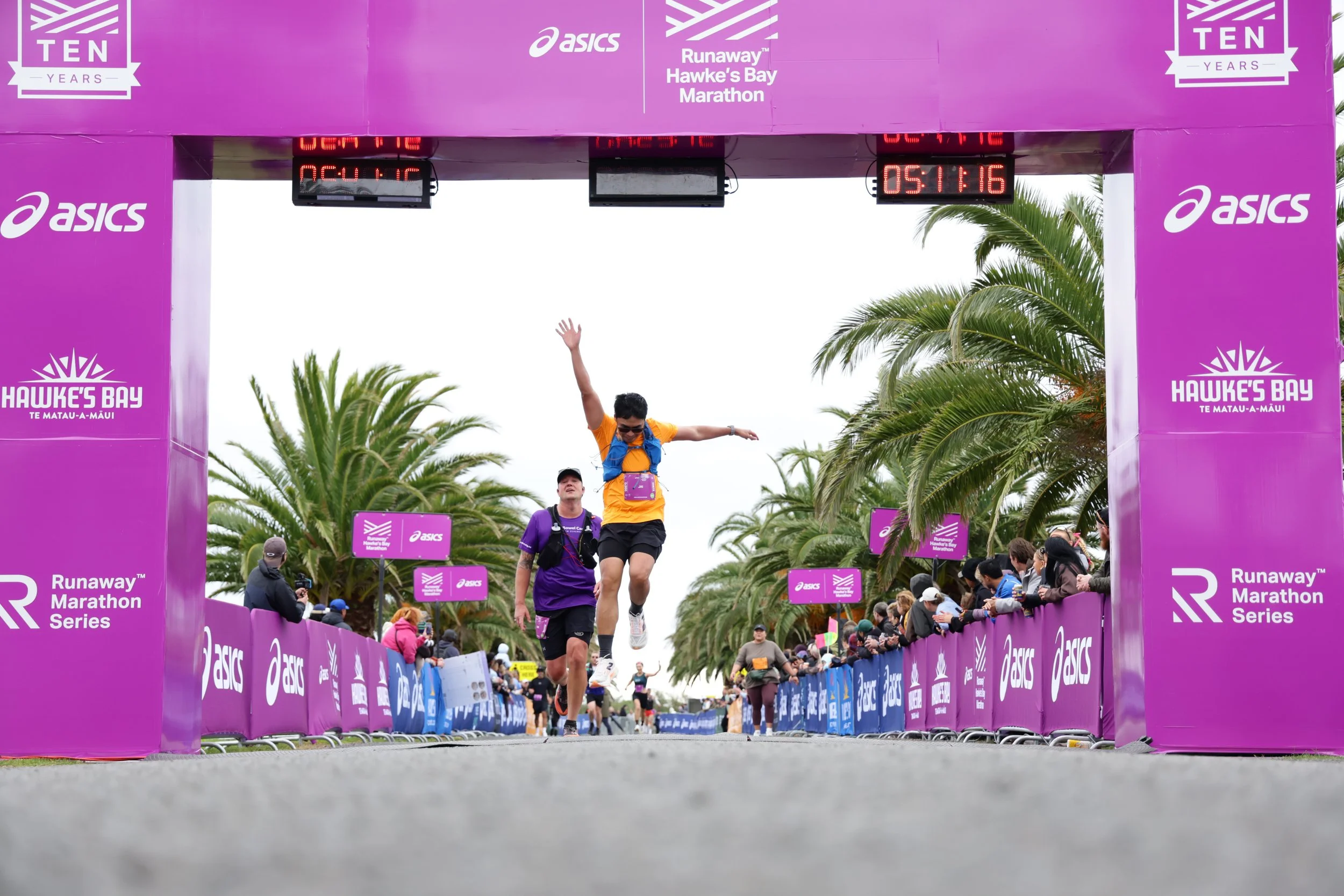Runner crossing the finish line at the Hawke's Bay Marathon event, with a crowd of spectators on either side, decorated with purple banners and palm trees in the background.