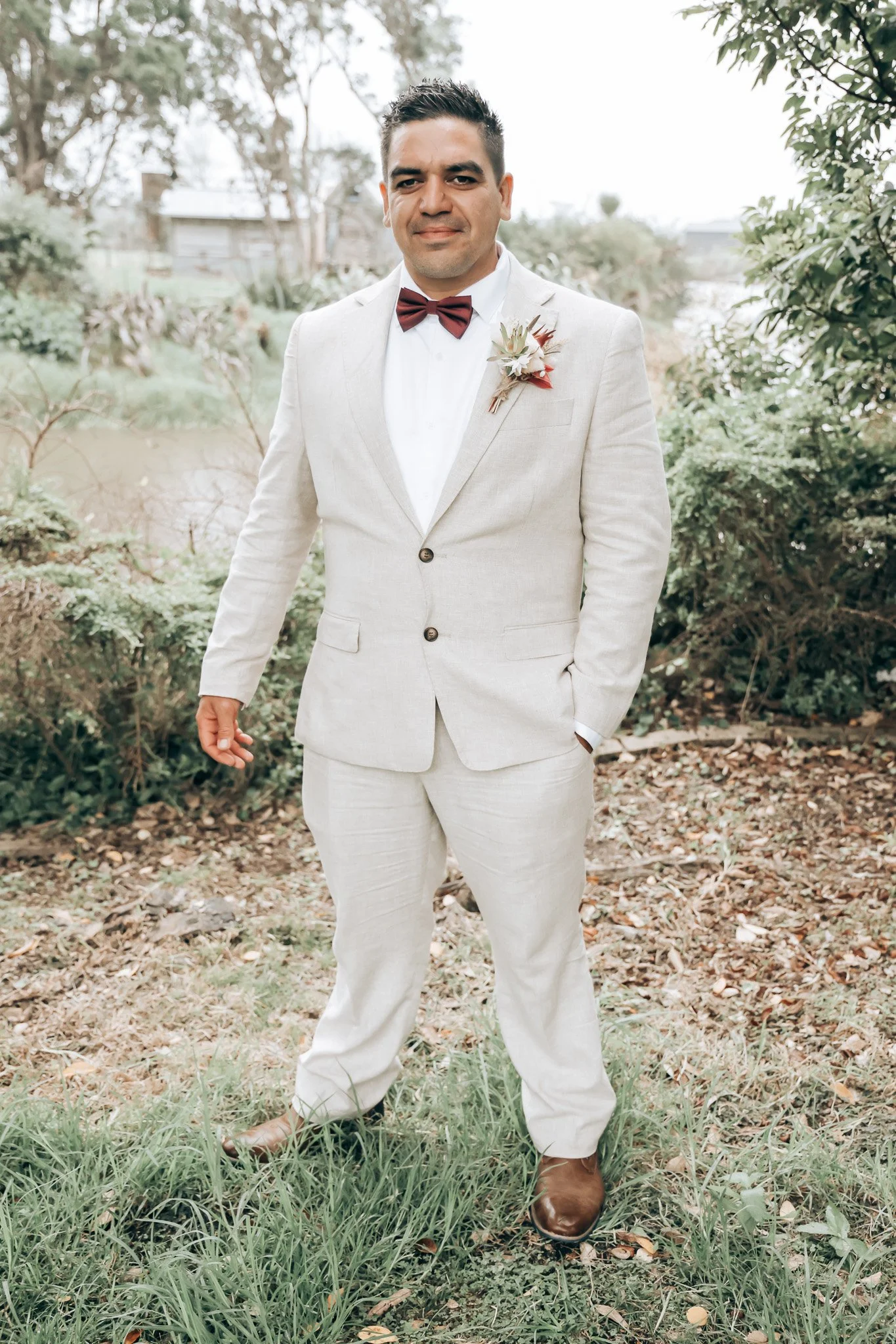 A man dressed in a light-colored suit with a white shirt, maroon bow tie, and a boutonnière, standing outdoors in a garden area with trees, bushes, and grass.