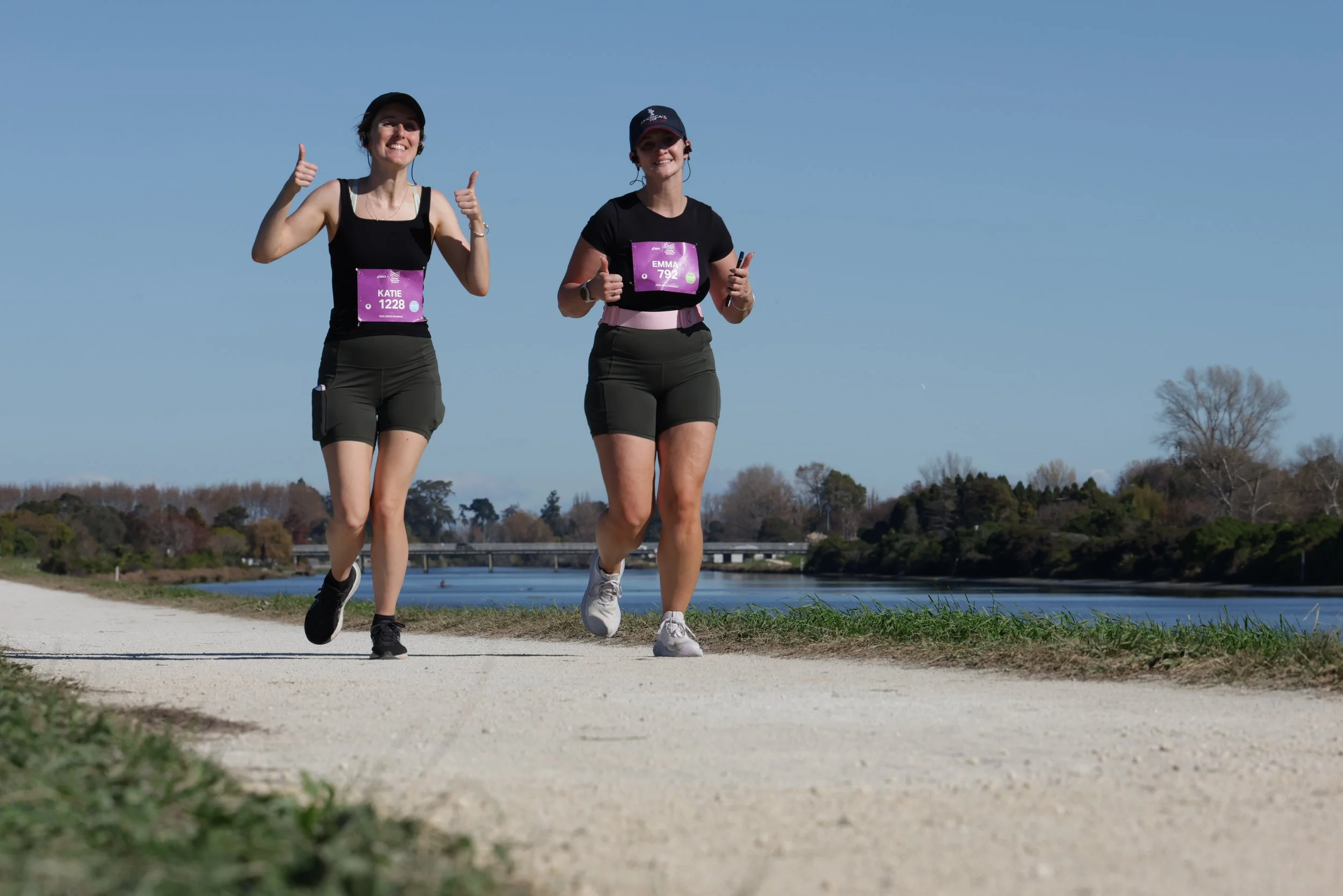 Two women running outdoors on a dirt path near a body of water with trees and a bridge in the background, smiling and giving thumbs up.