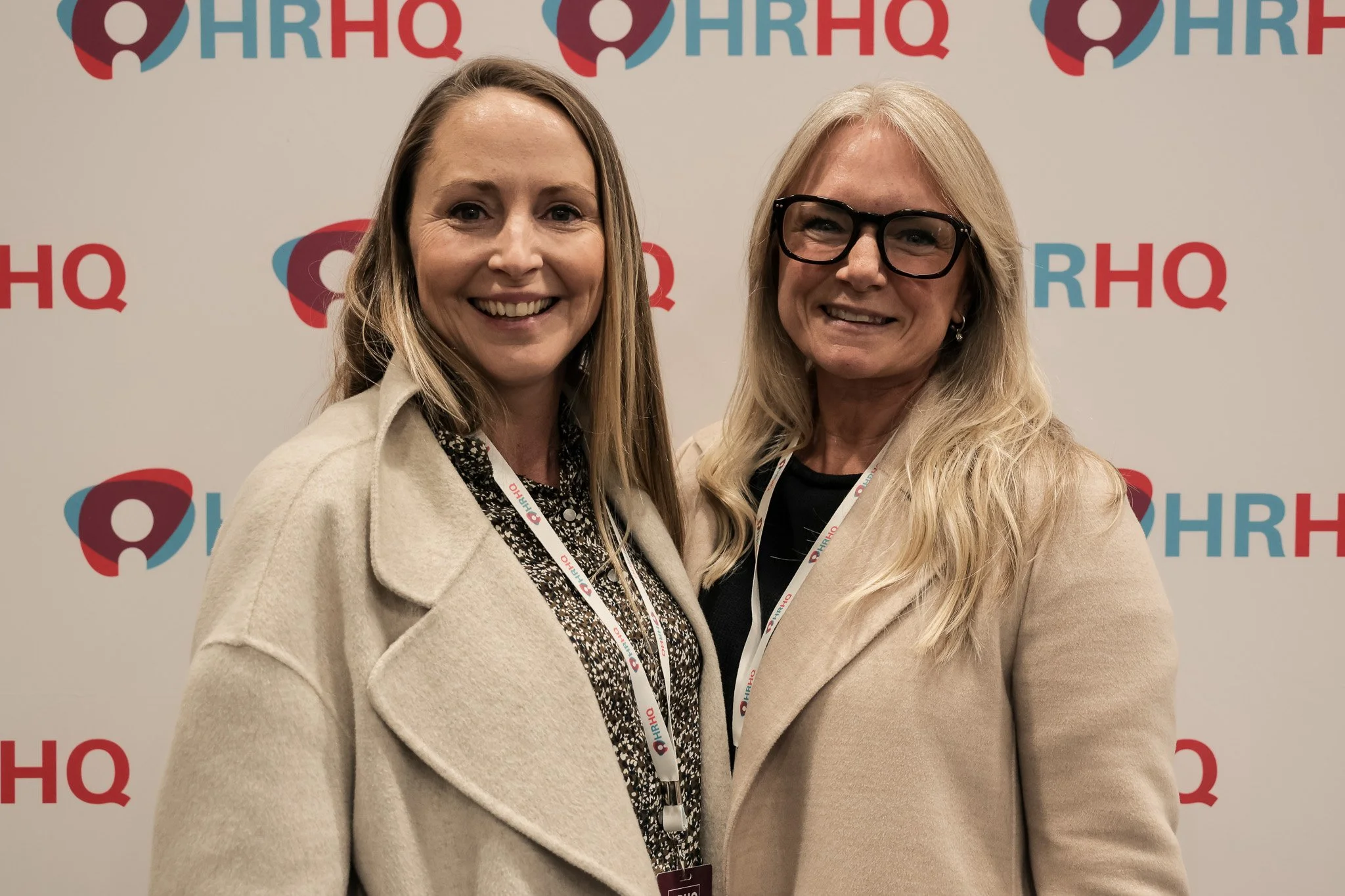 Two women standing close together smiling in front of a white background with a red and blue logo that reads 'HRHQ' repeatedly.