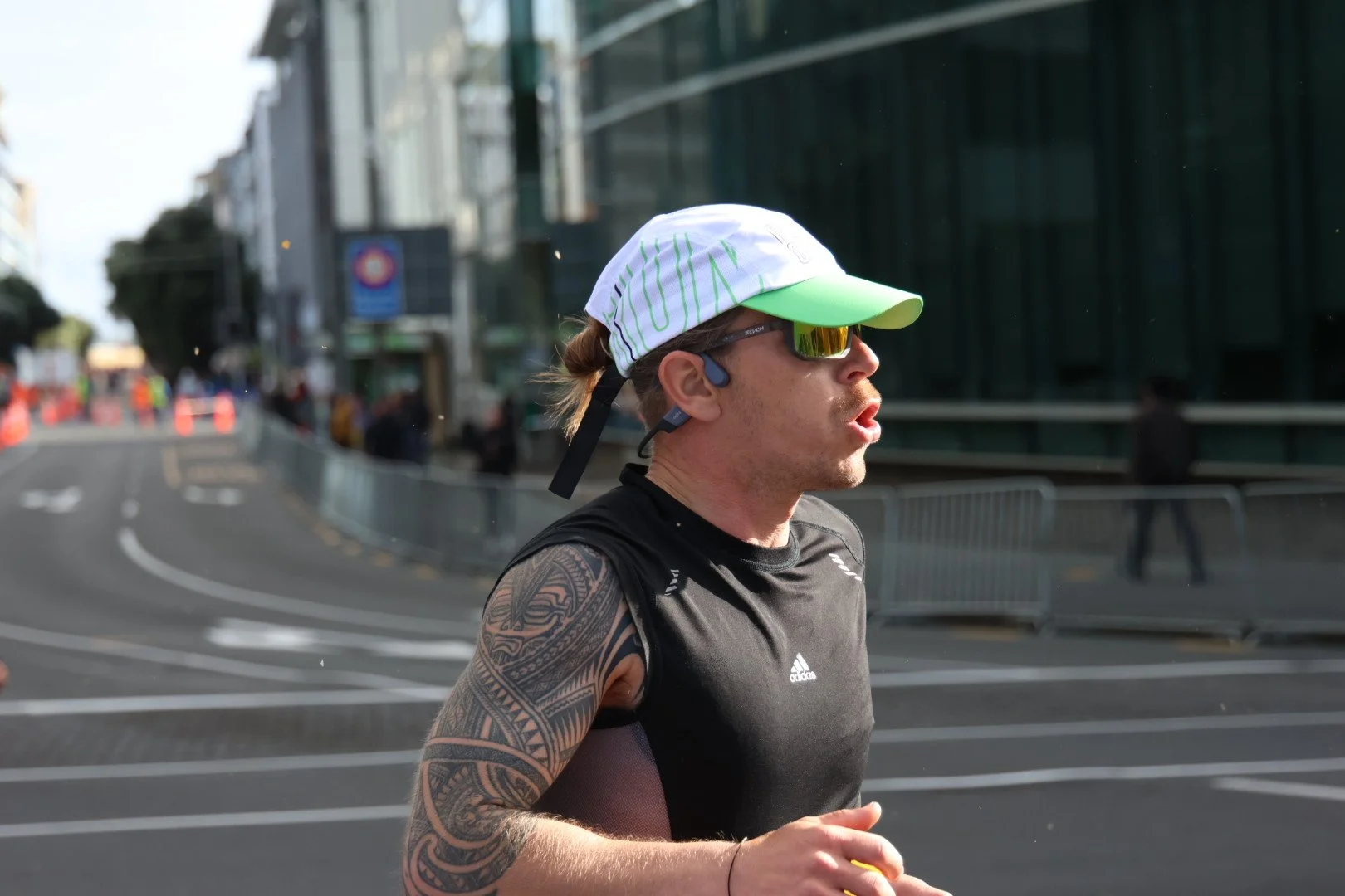 Male runner in black athletic gear with a tattooed arm, white cap, and sunglasses, running past urban buildings and barriers during daytime.