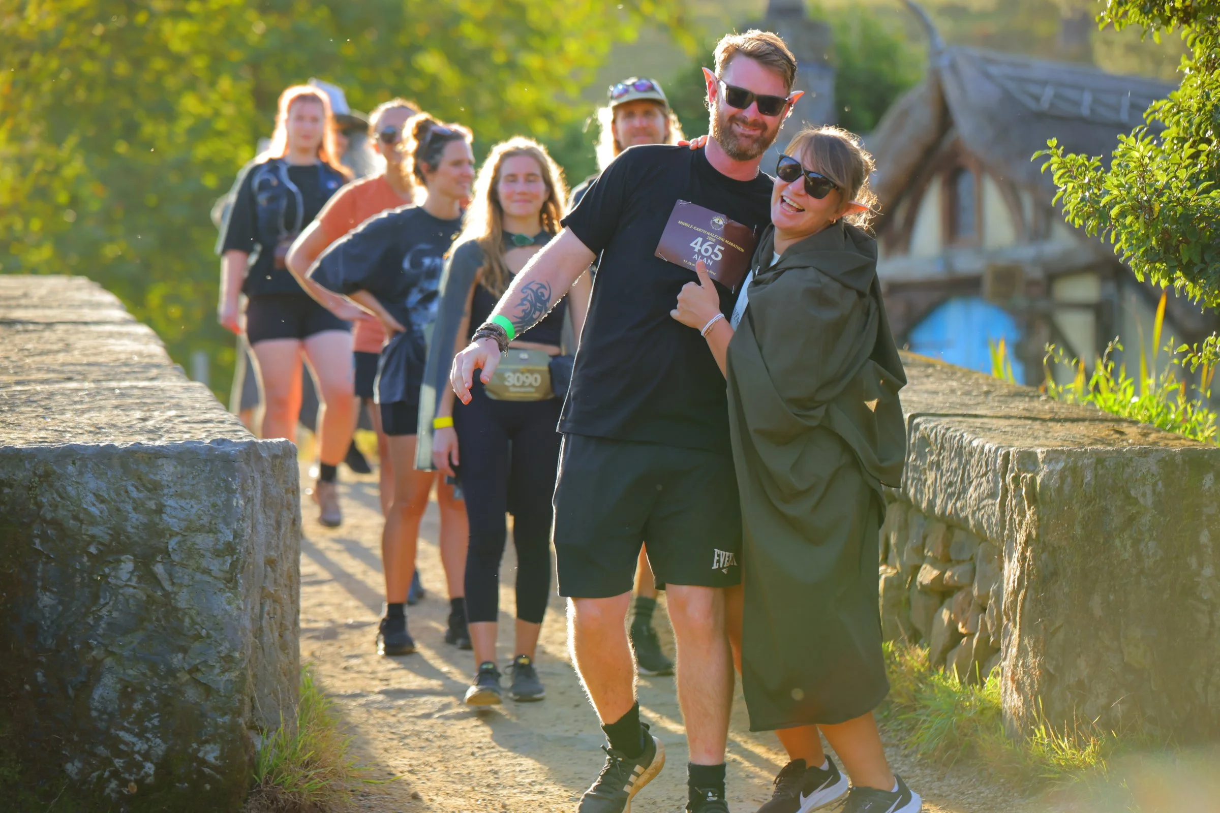 A group of people dressed in athletic gear, standing in a line along a stone pathway outdoors in a park, with two of them in the foreground smiling, hugging, and wearing sunglasses. The background is filled with green trees, and the scene is bathed i
