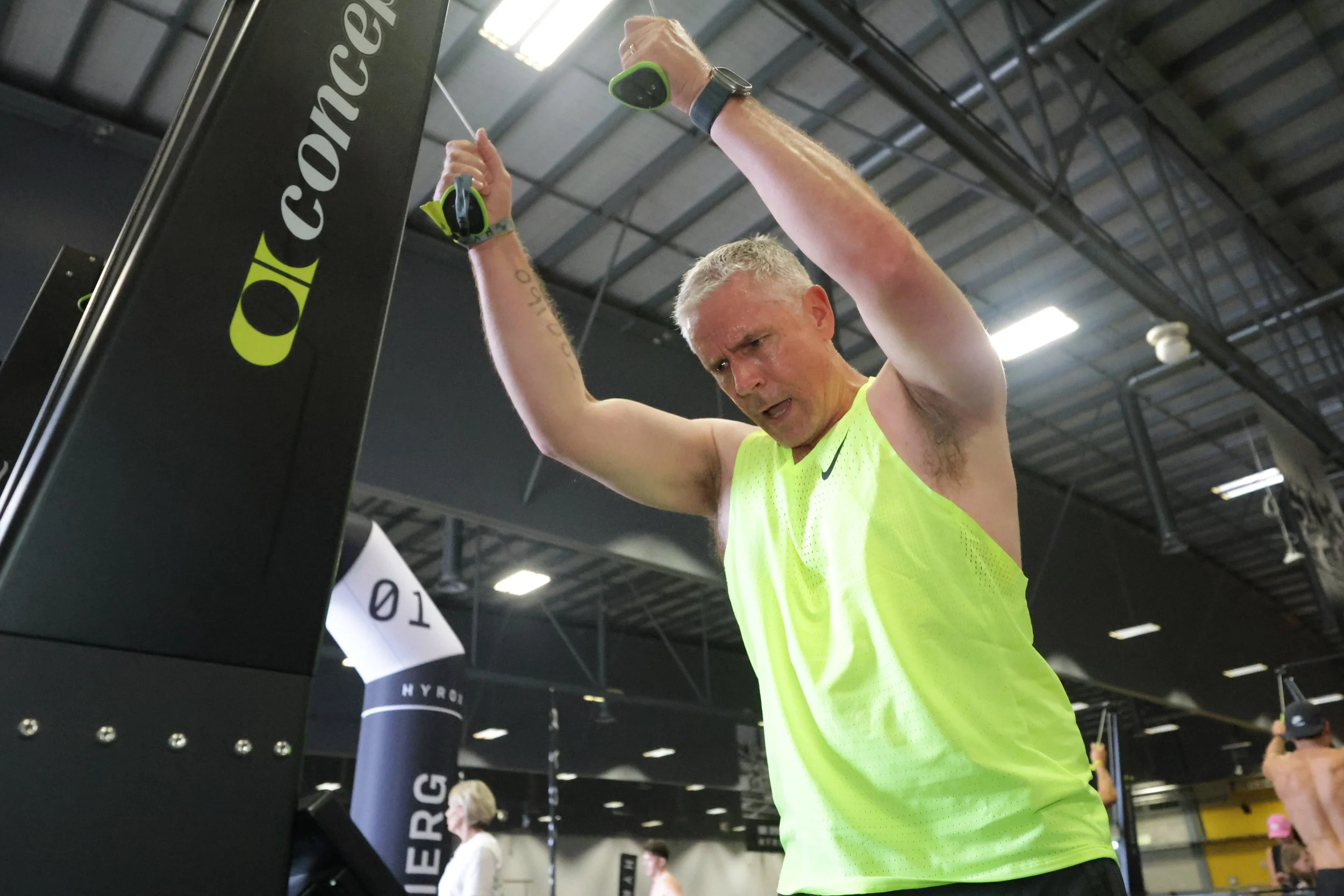 A middle-aged man with short gray hair wearing a safety watch and a neon yellow sleeveless shirt works out at a fitness gym, pulling on a resistance band attached to a black workout station.