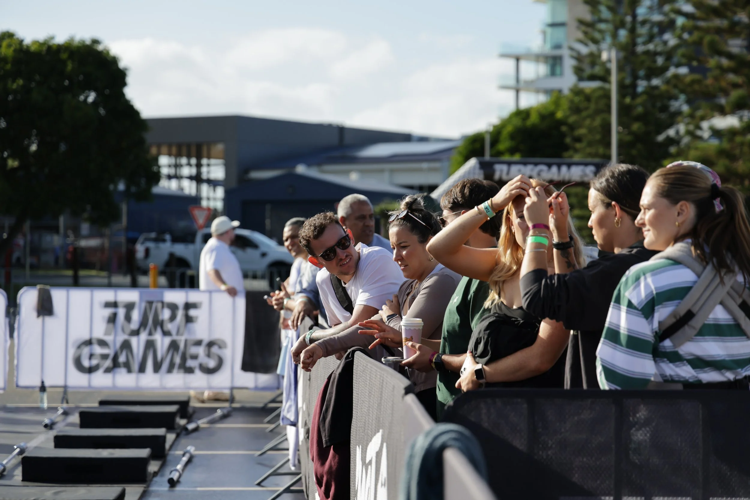 Group of people sitting and standing behind a barrier at an outdoor event, with some smiling and talking, during daytime with a partly cloudy sky.