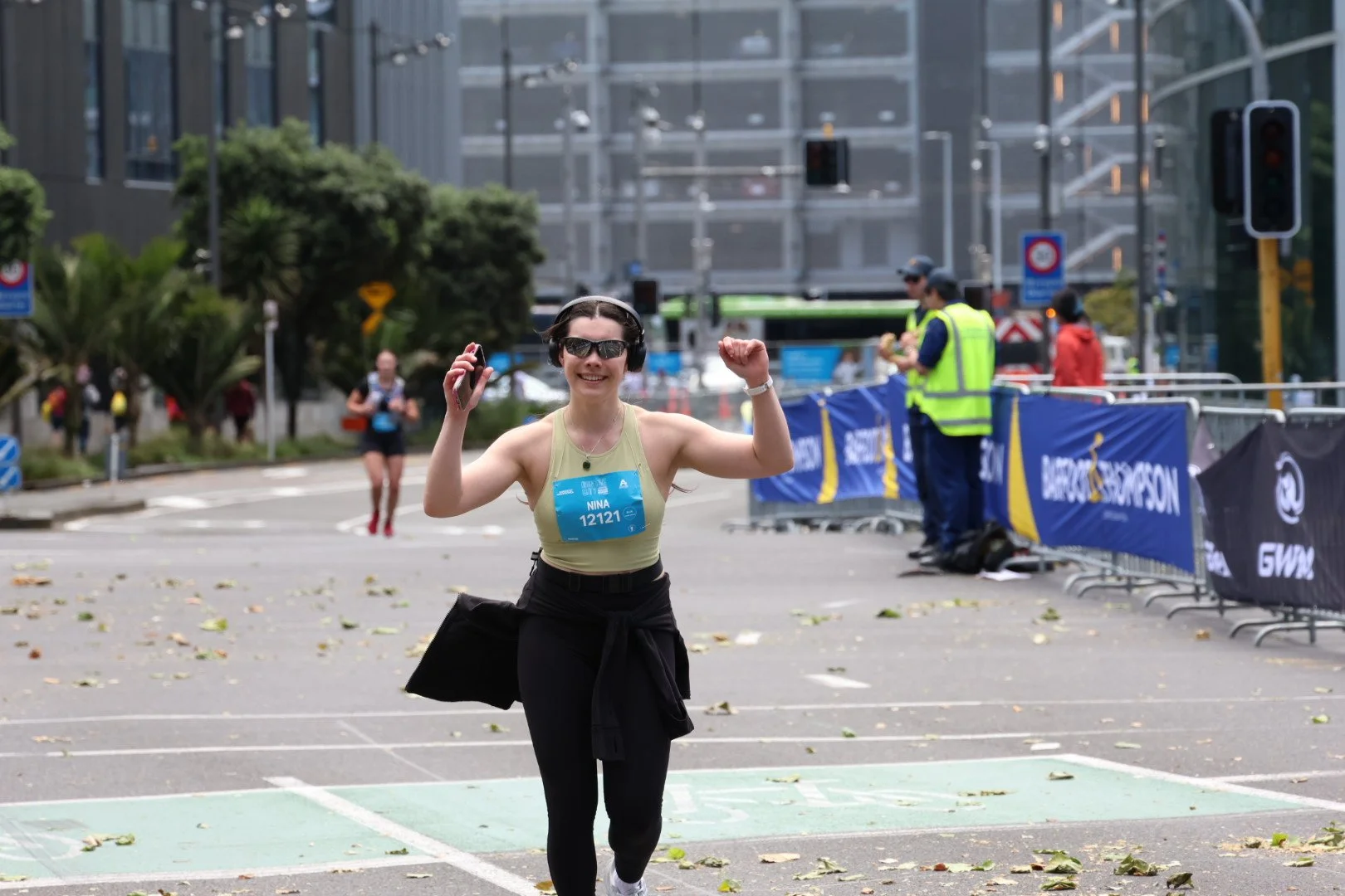 A woman in athletic wear, sunglasses, and headphones celebrates crossing the finish line of a race in an urban area.