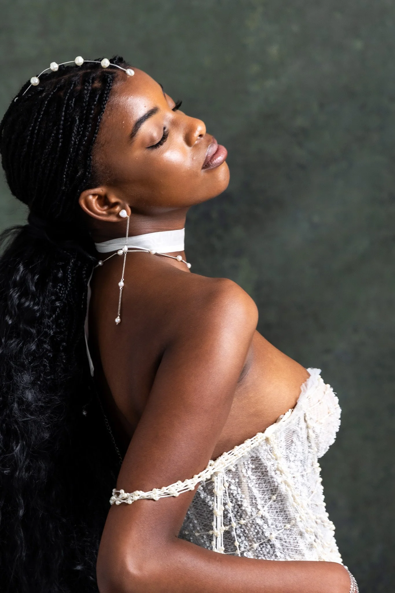 Profile of a woman with long braided hair, wearing pearl jewelry, and a white lace strapless dress, with eyes closed and a serene expression, against a dark background.