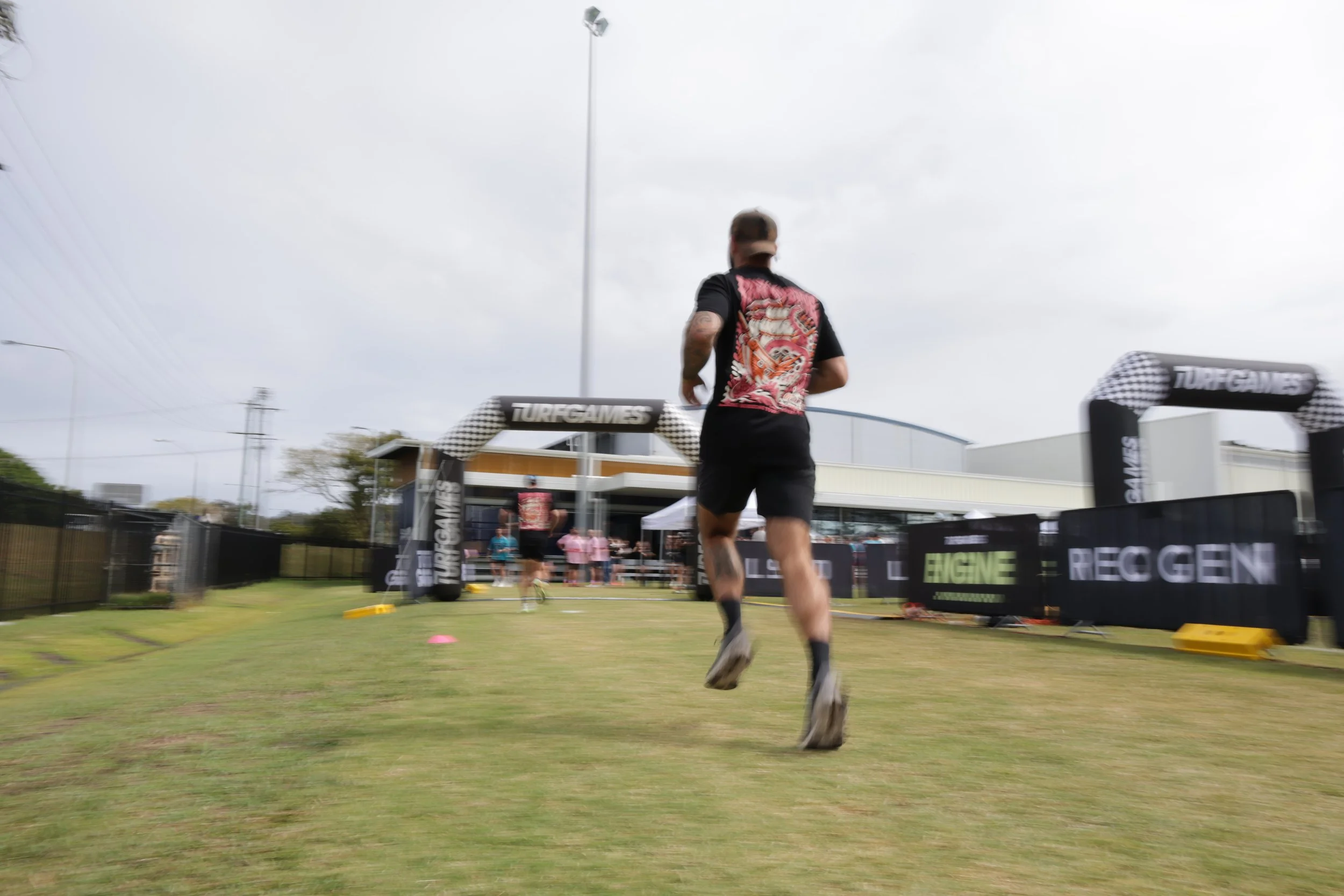 A man running in a race event outdoors with a finish line and spectators in the background on an overcast day.