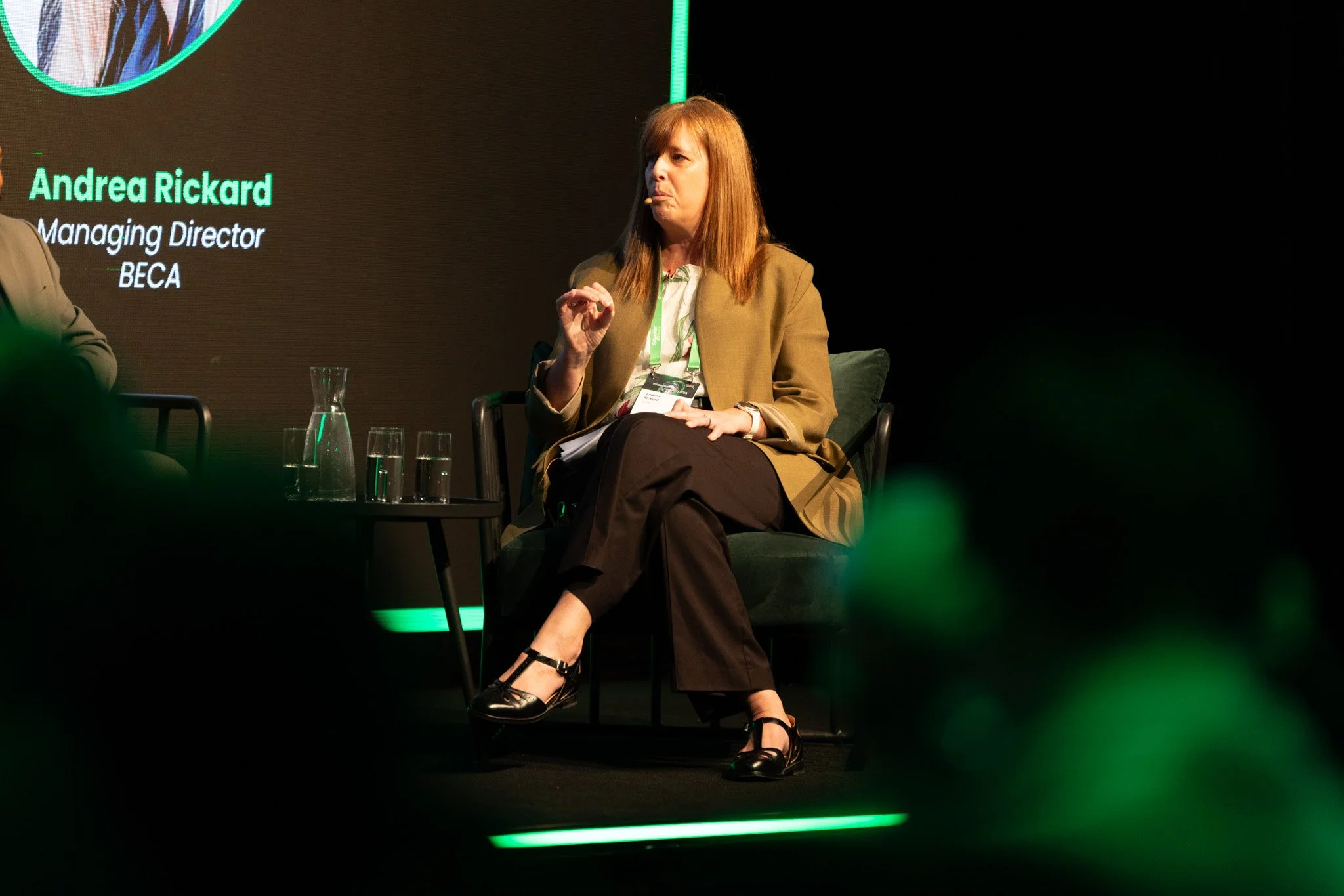 A woman with red hair wearing a tan blazer, white blouse, and black pants sitting on a chair on stage during a panel discussion, with a presentation slide and glasses of water on a small table nearby.