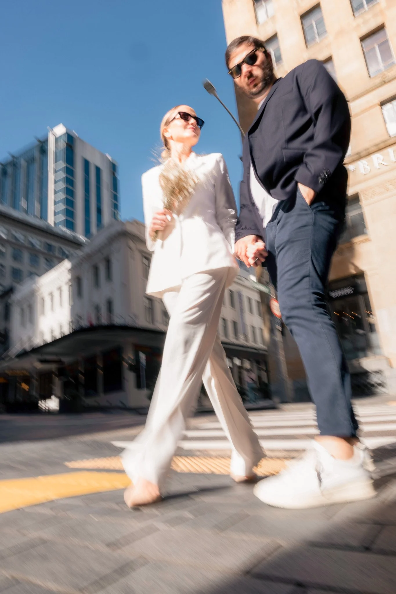 Two people crossing the street in an urban area, wearing stylish and casual clothes, with modern buildings in the background under a clear blue sky.