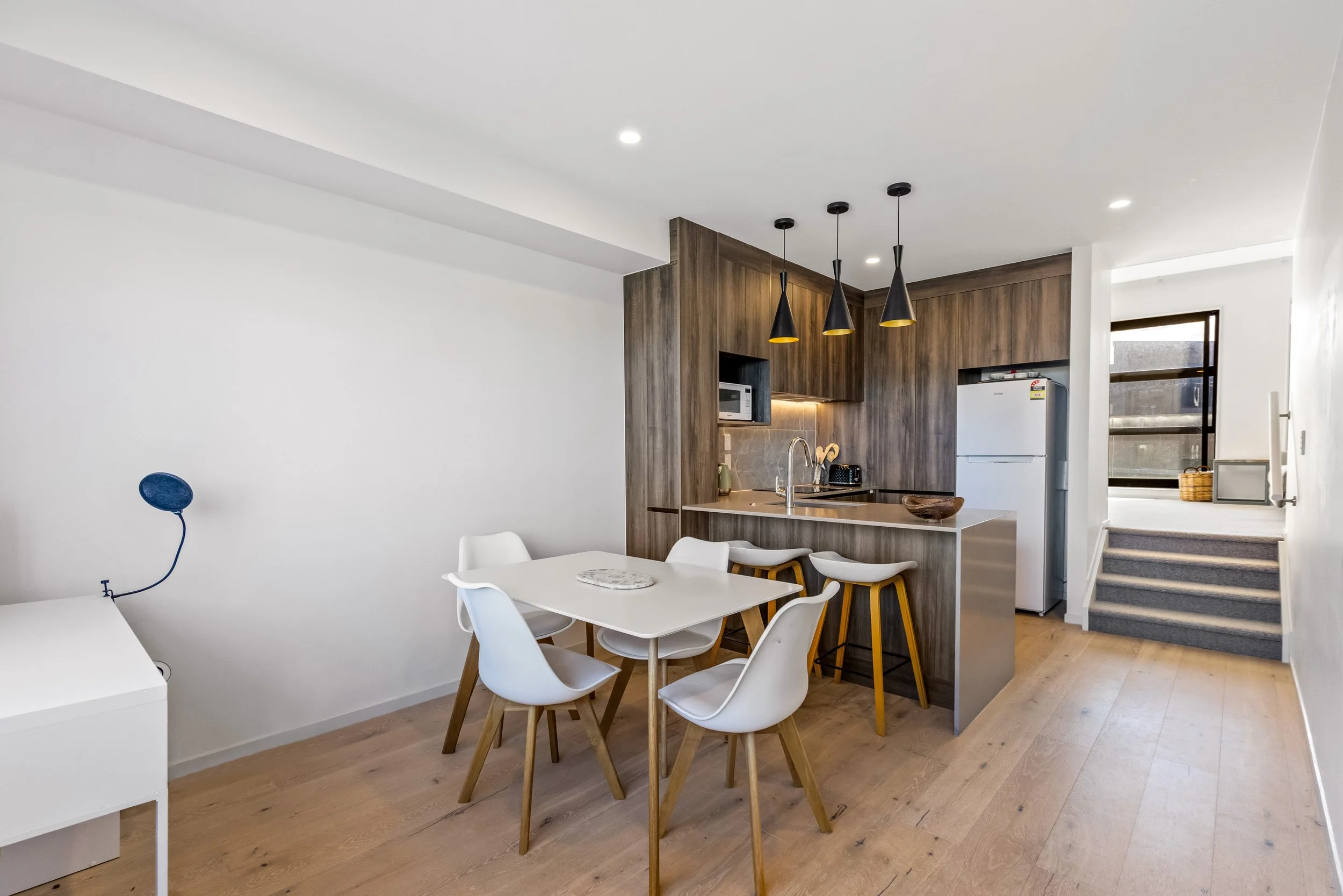 Modern kitchen with wooden cabinets, white walls, black pendant lights, white dining table with white chairs, and stairs leading upstairs.