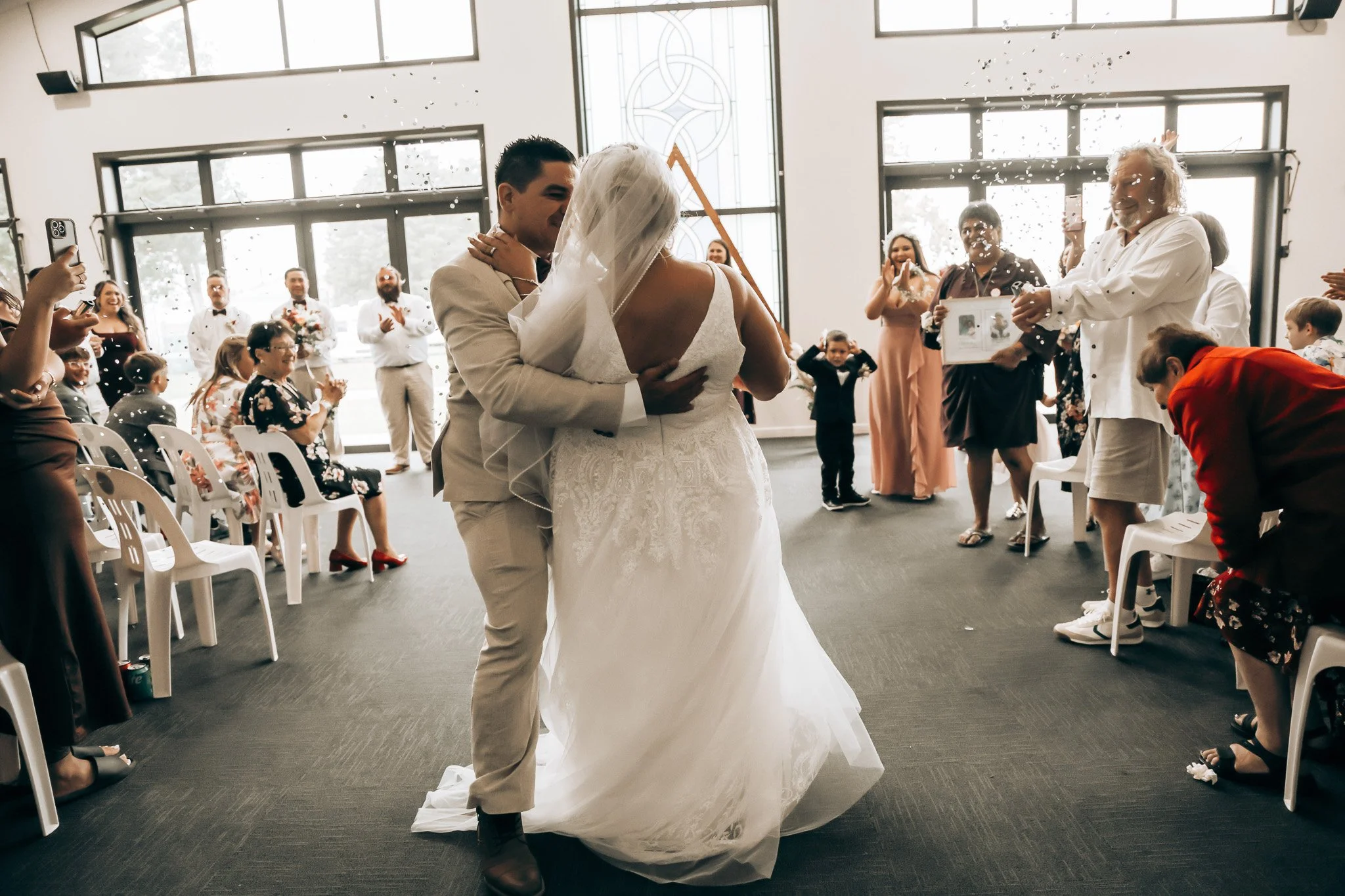 A couple, dressed in wedding attire, is dancing closely while guests gather around in a spacious, brightly lit room with large windows and modern decor. The bride is in a white gown with a veil, and the groom is in a light-colored suit.