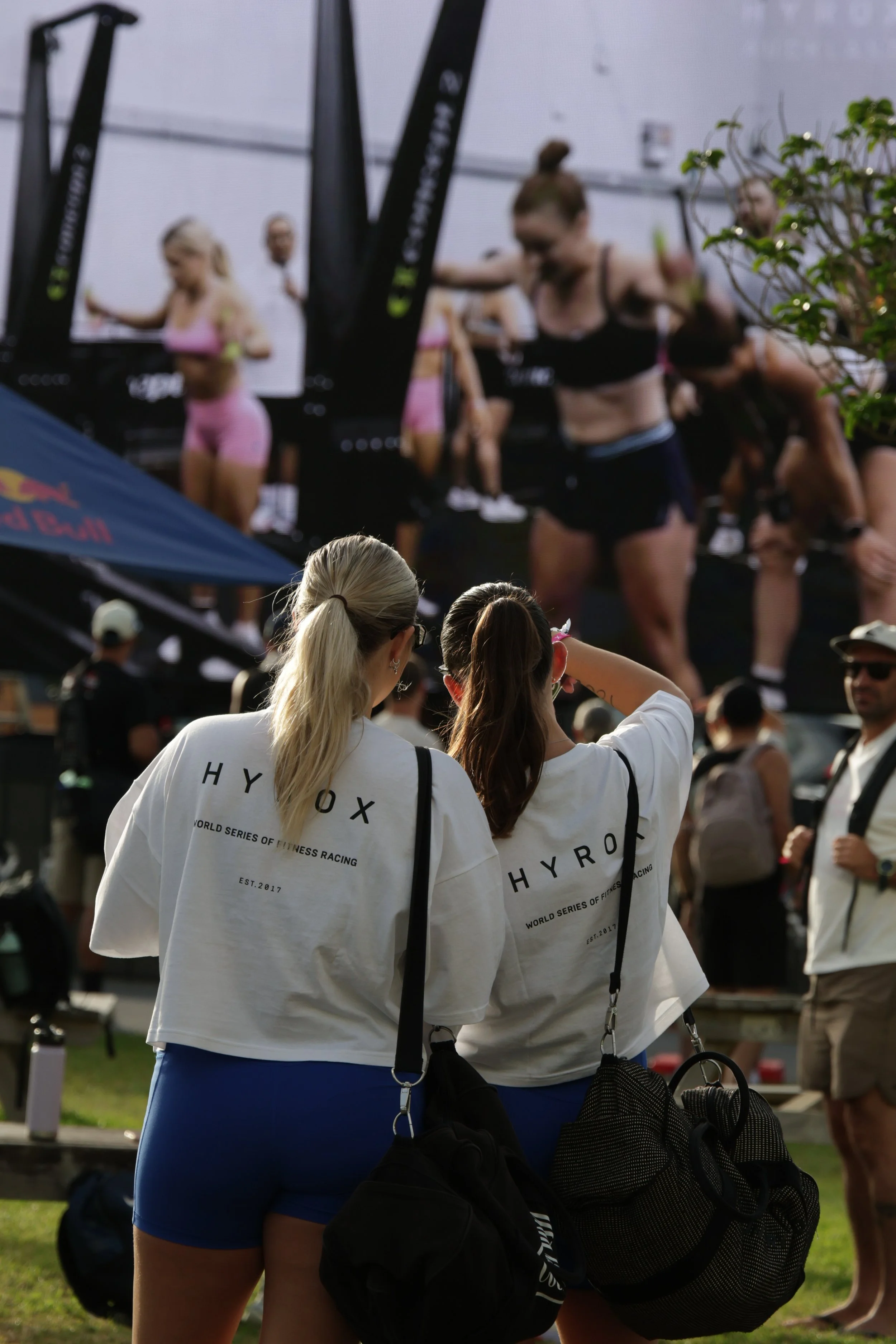 People at a fitness event, with women seen from behind wearing white t-shirts with 'HYROX' printed on them, and others on a stage or platform in athletic wear.