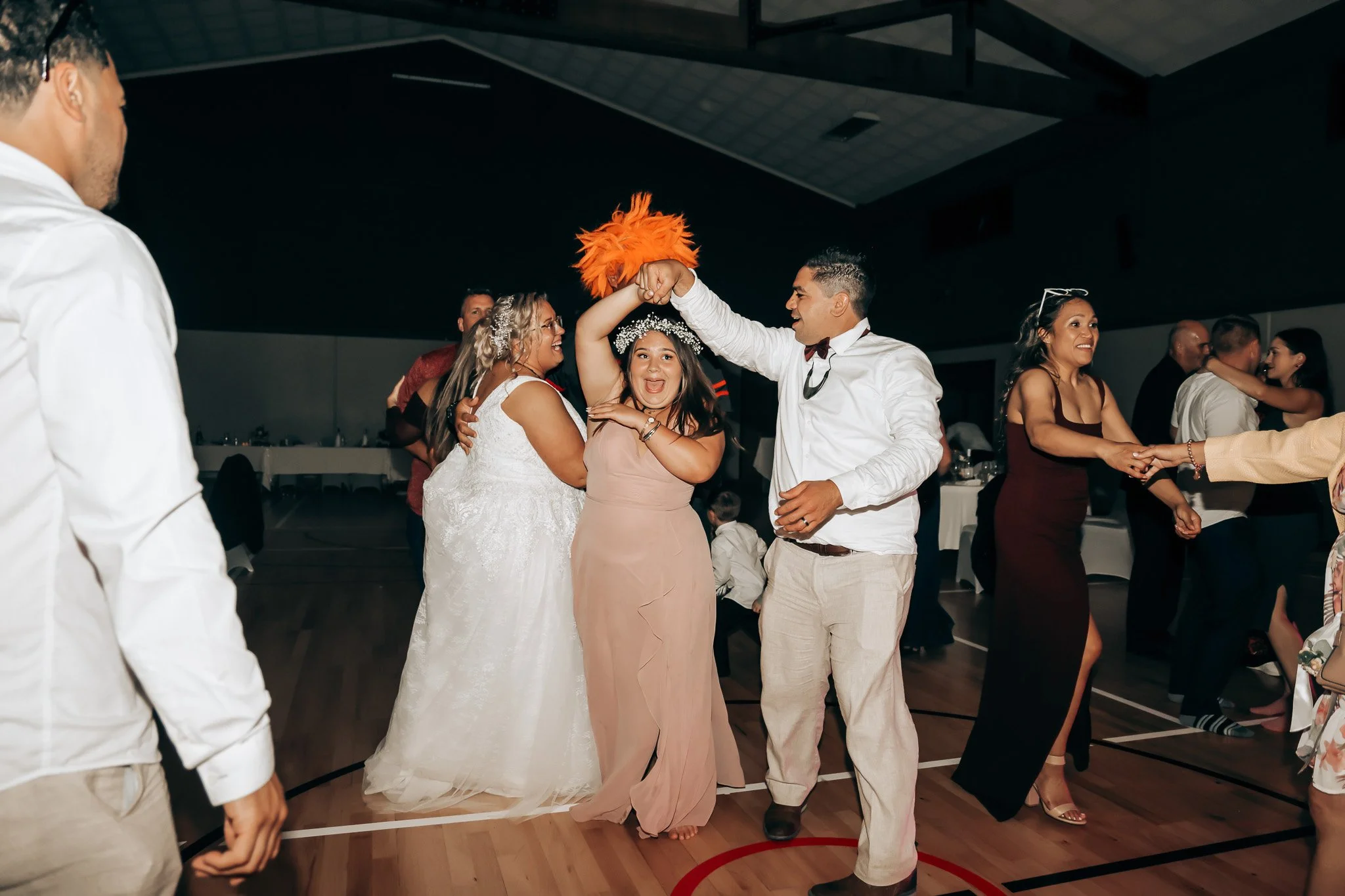 People dancing and celebrating at a wedding reception indoors, with a man in a white shirt and bowtie and a woman in a wedding dress, surrounded by friends and family.