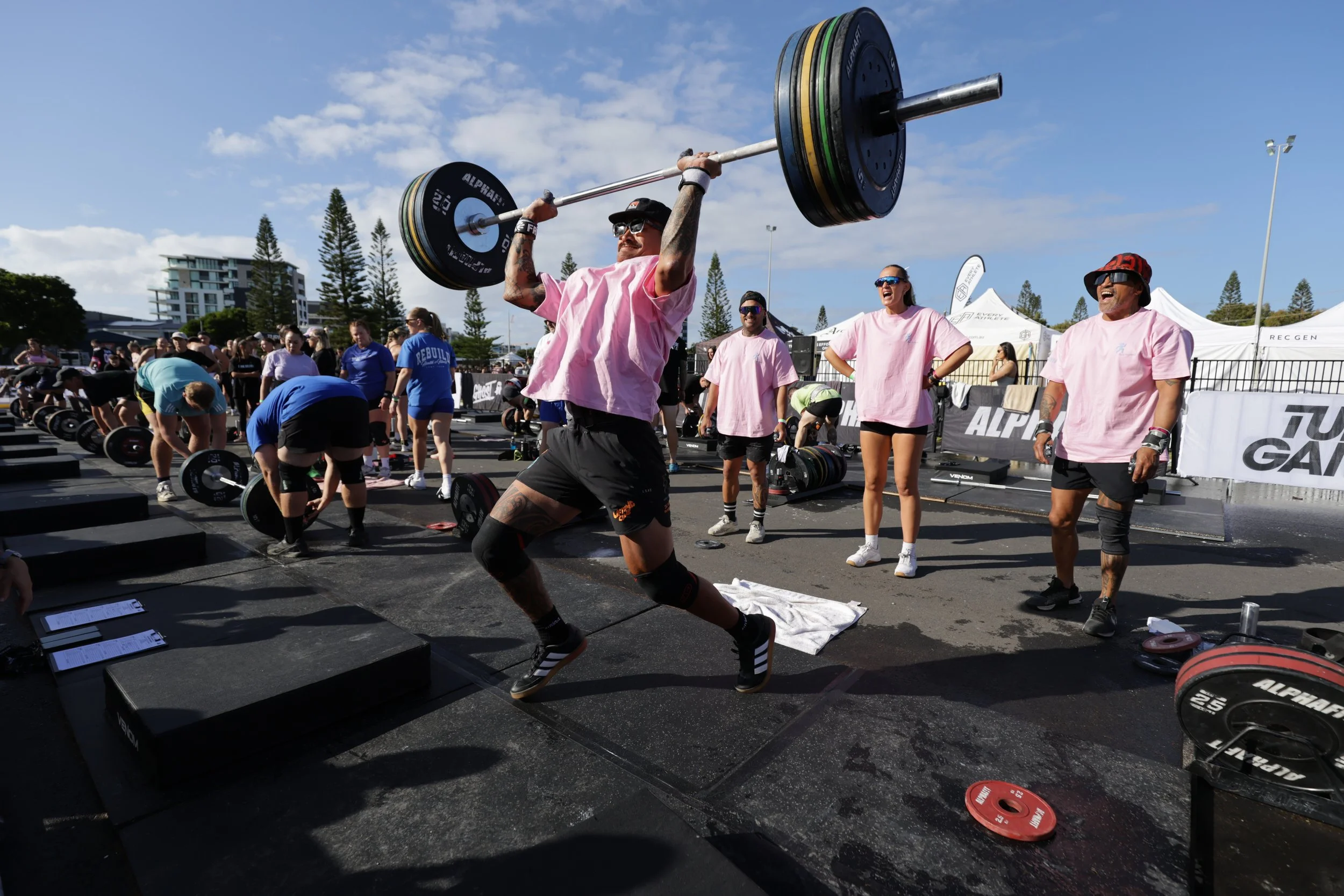 A man in a pink shirt lifts a barbell overhead during an outdoor fitness event, with several onlookers standing or working out nearby, tents and trees in the background, and a partly cloudy sky overhead.