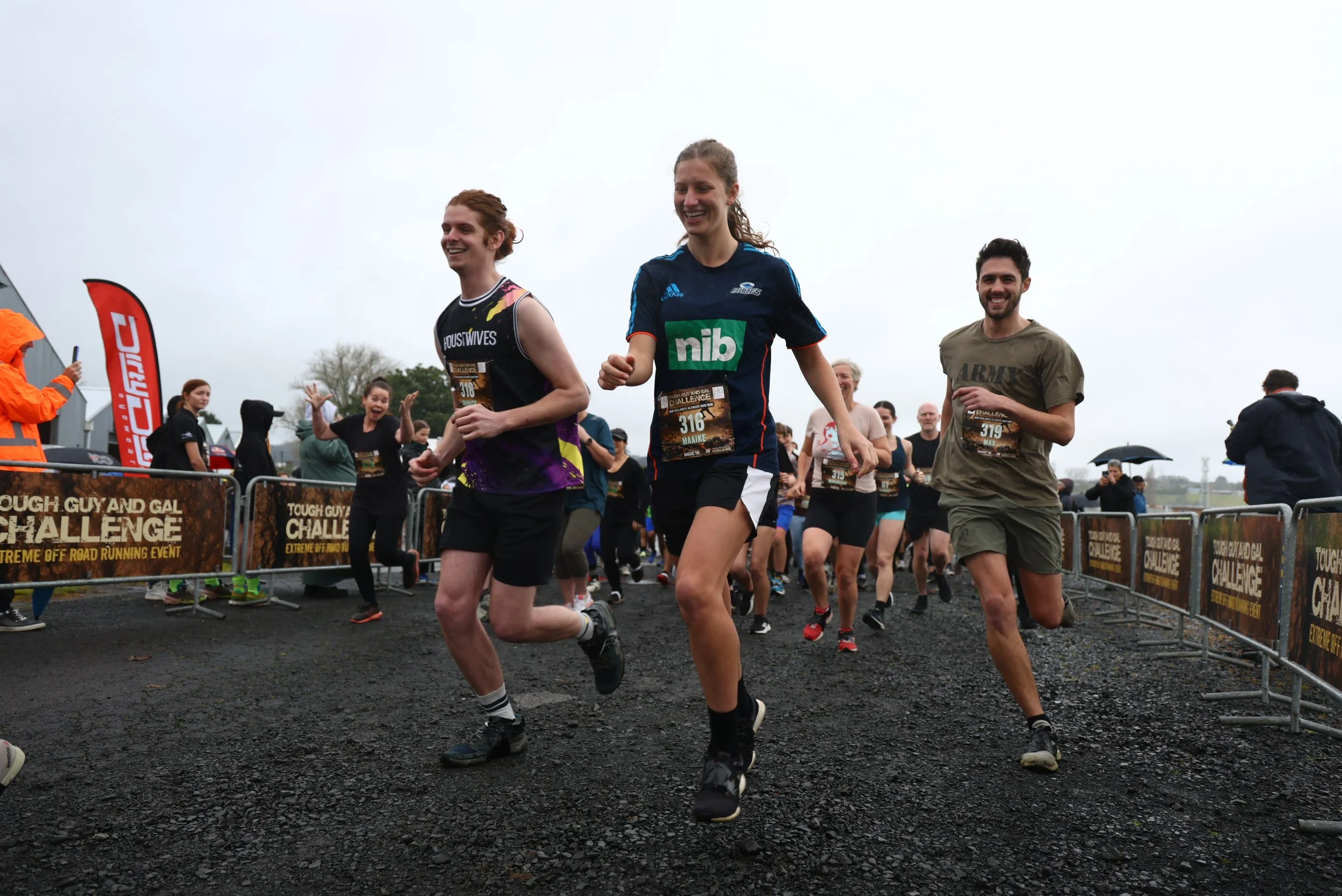 Group of runners participating in an outdoor race, with some smiling and others focused, under a cloudy sky, with spectators behind a barrier.