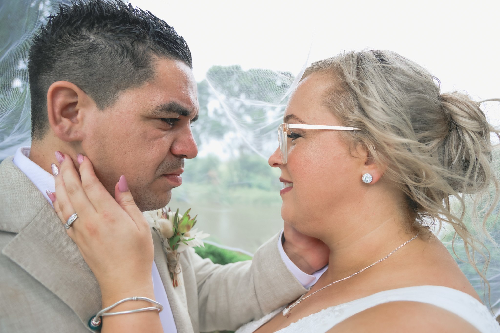 A close-up of a couple on their wedding day, gazing into each other's eyes, with the bride touching the groom's face gently.