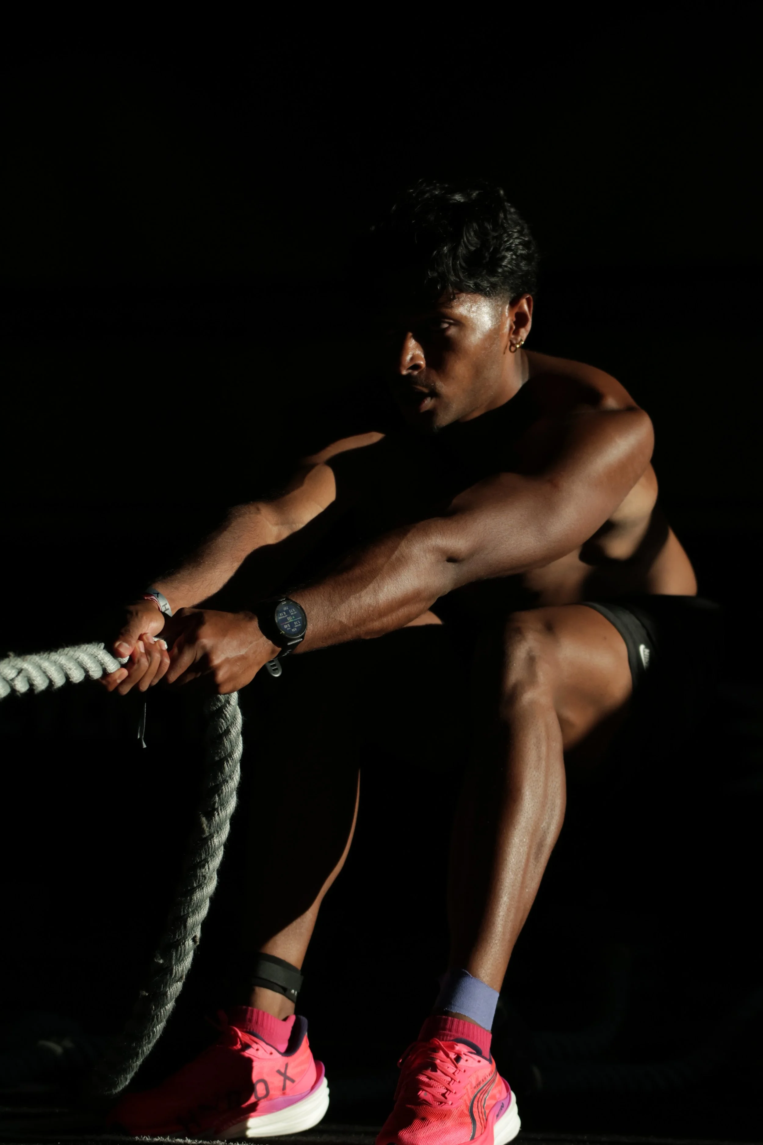 A person with dark, curly hair engages in a workout, holding a thick jumping rope and wearing pink athletic shoes against a dark background.