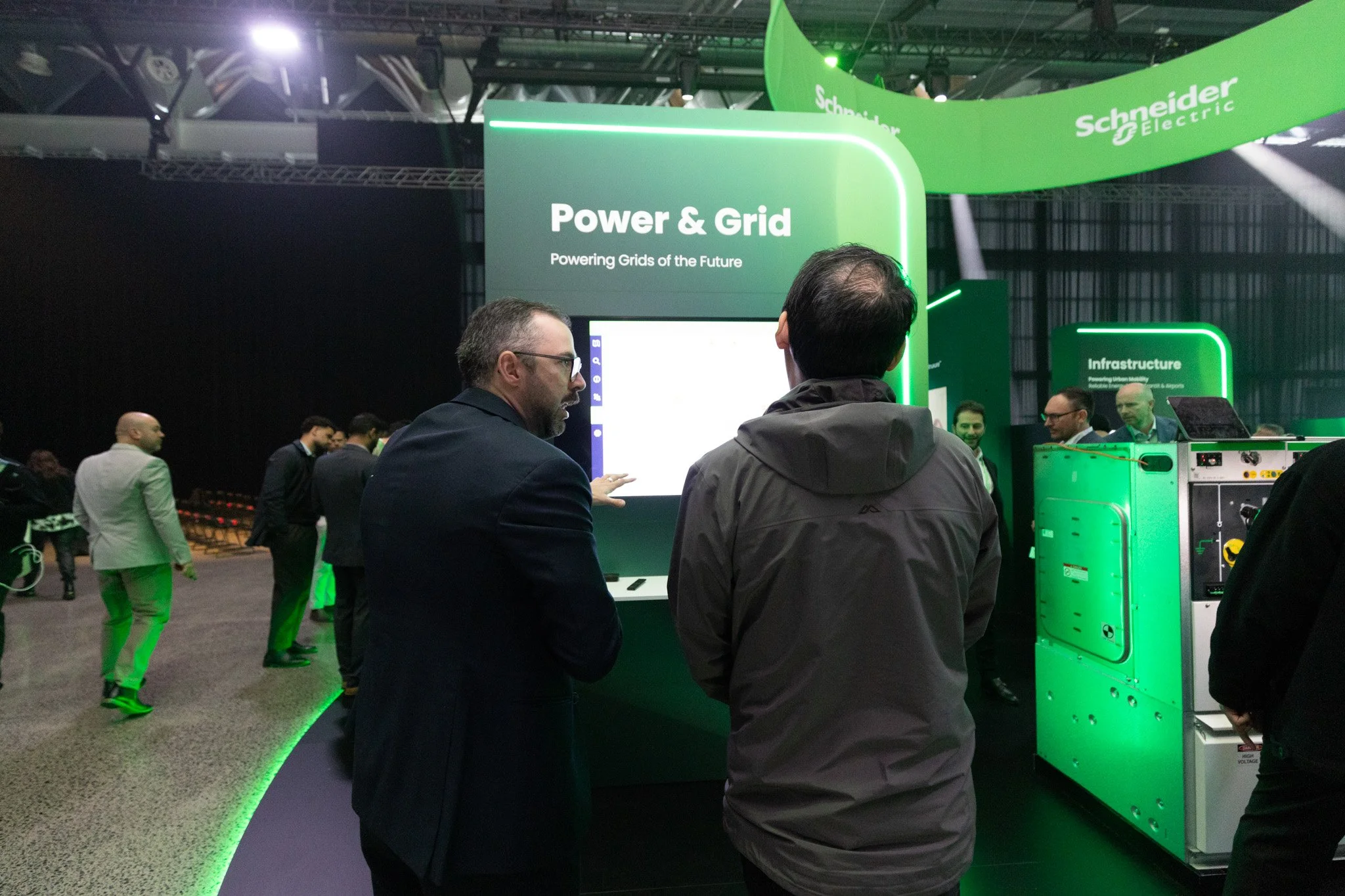 At a trade show, attendees are gathered around a booth for Schneider Electric with a sign reading 'Power & Grid' and 'Powering Grids of the Future.' The booth features green lighting and equipment related to electrical power and grid infrastructure. 