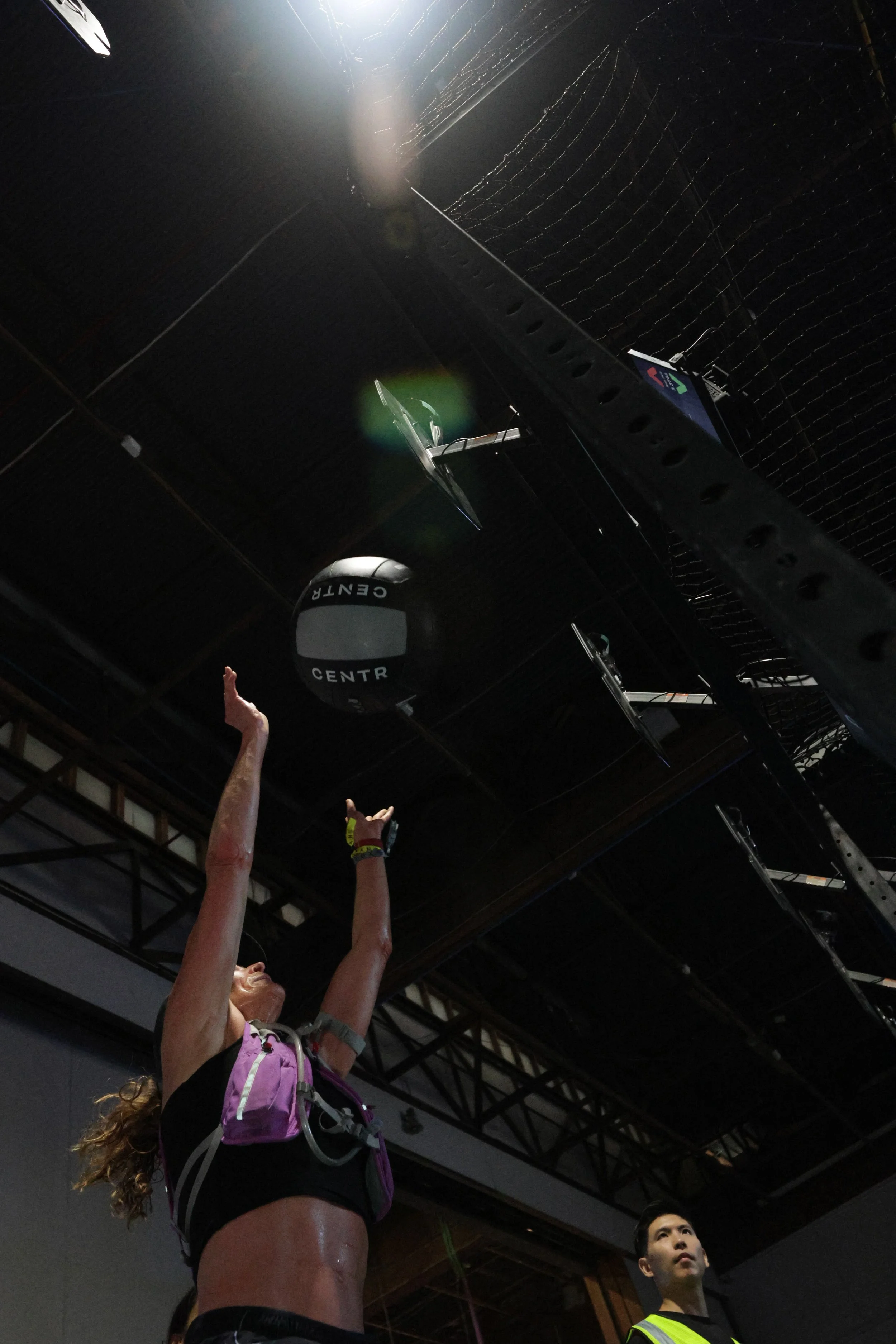 A woman in athletic gear reaching upward with her arms raised inside a gym or sports facility. In the background, a man in a reflective vest is looking upward. The ceiling has black panels, lighting, and equipment attached.