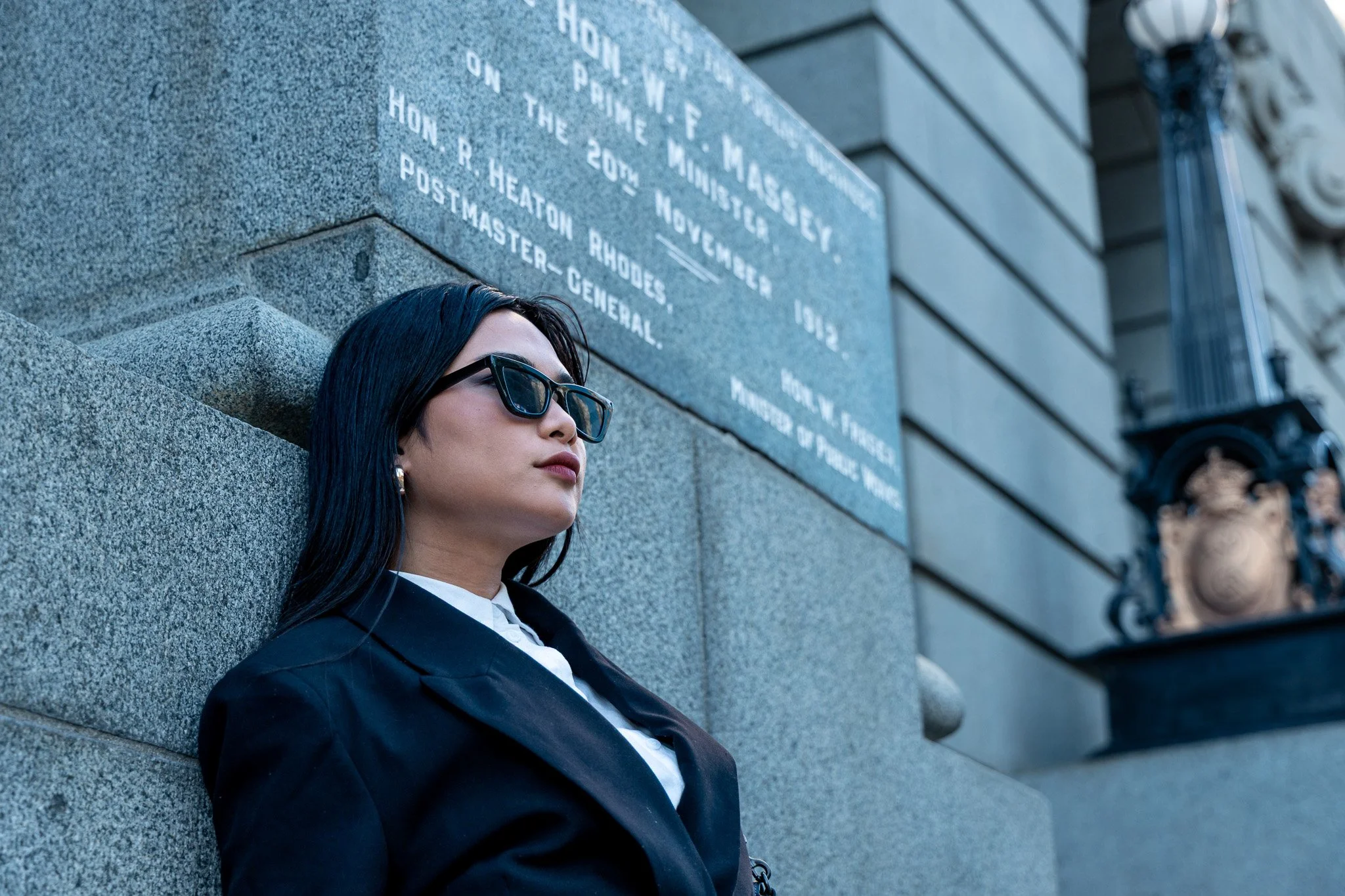 A woman with long dark hair, wearing a black suit, white shirt, and sunglasses, leaning against a stone wall with engraved text, in an urban setting.