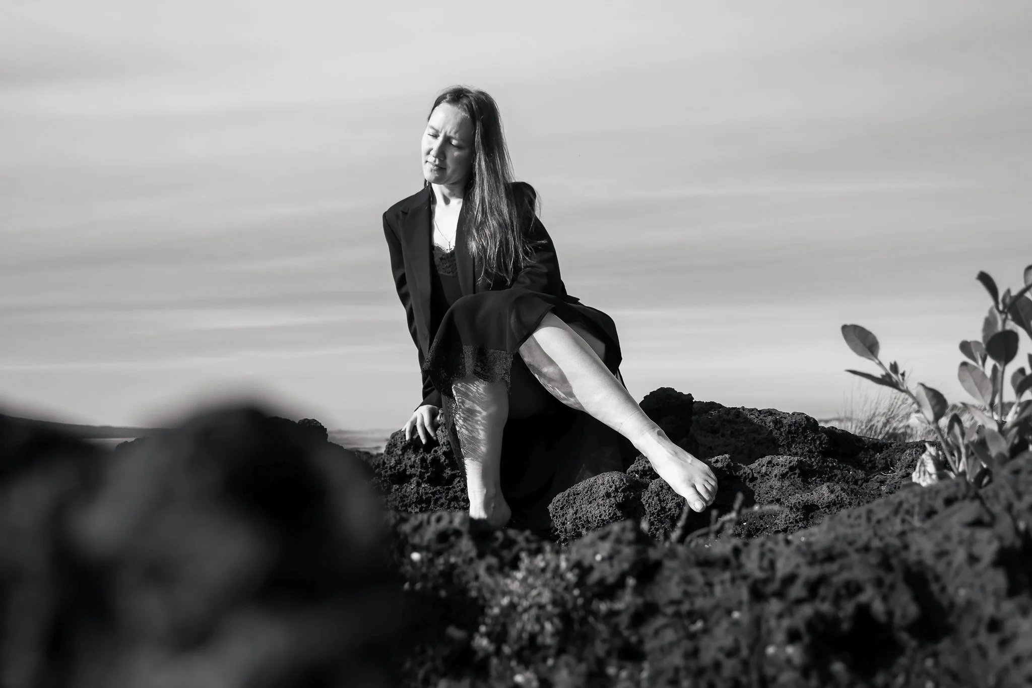 A black and white photo of a woman sitting on rocky terrain with her eyes closed, wearing a dark blazer over a lace top. The sky is cloudy, and some foliage is visible in the foreground on the right.