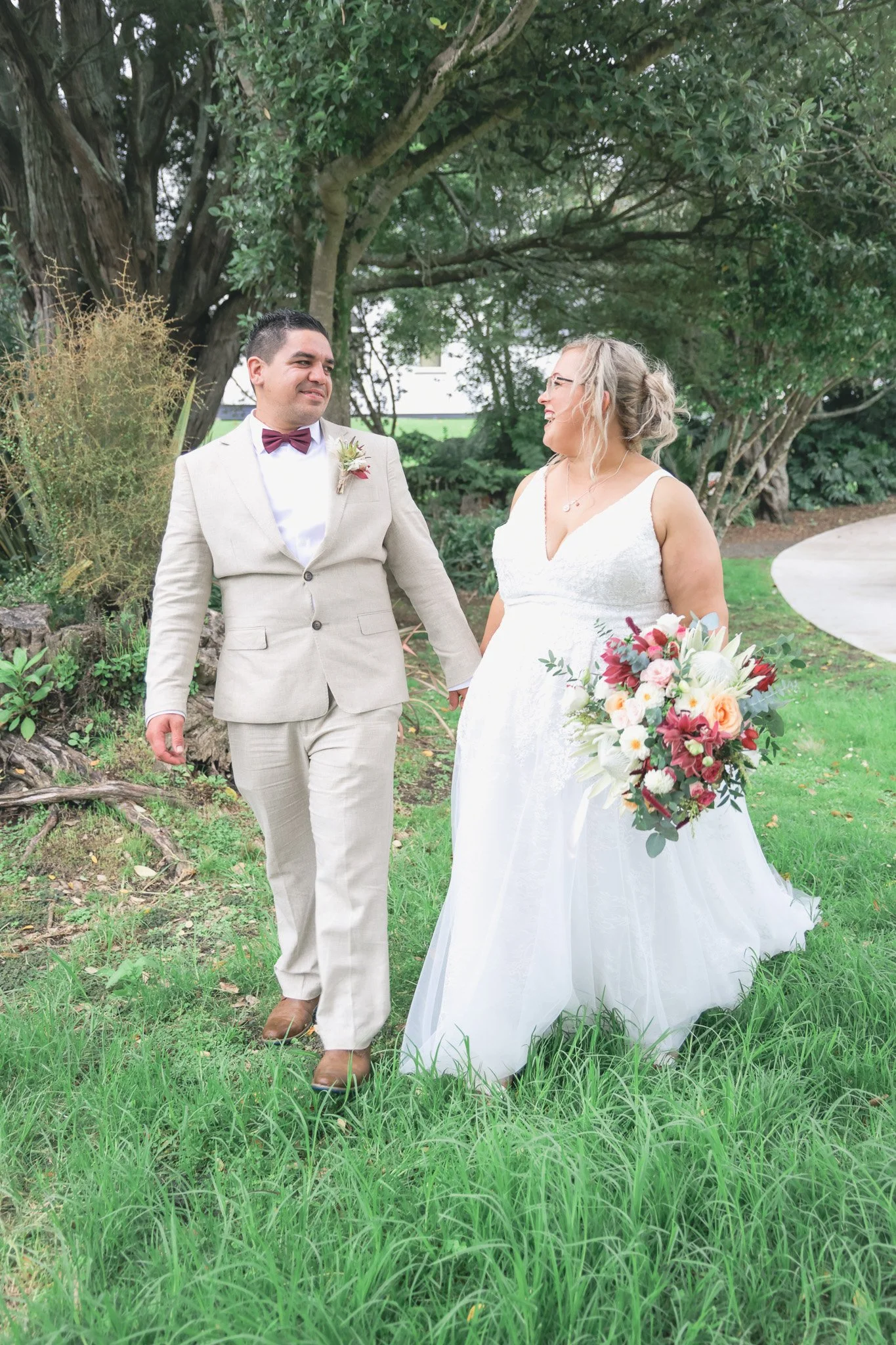 A bride and groom holding hands and walking outdoors on a grassy pathway, surrounded by trees and greenery, during a wedding ceremony.