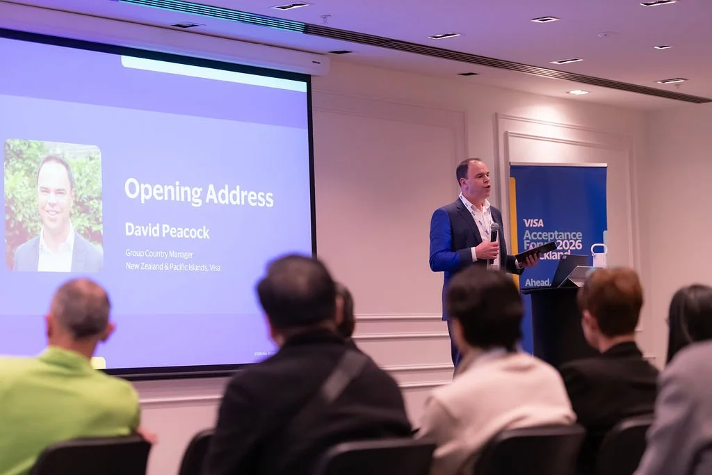 A man in a suit standing next to a podium, speaking to an audience at a conference, with a presentation slide on the screen behind him. The slide displays the text 'Opening Address,' 'David Peacock,' and his title as Group Country Manager, New Zealan