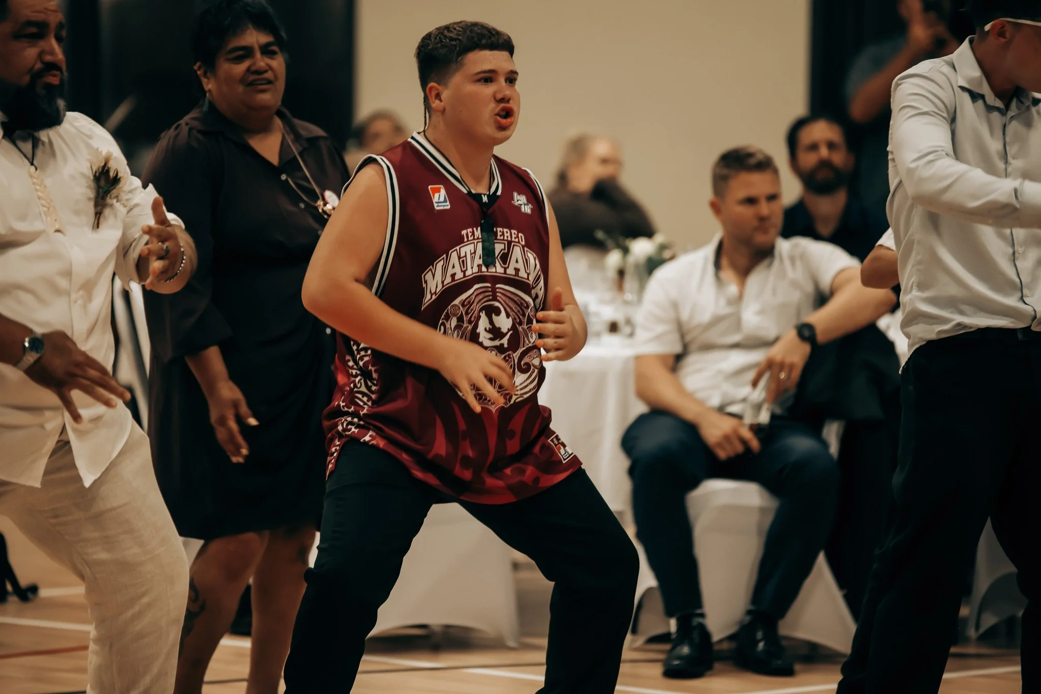 Young man in a maroon basketball jersey participating in a group dance or activity, surrounded by diverse seated and standing people in a well-lit indoor setting.