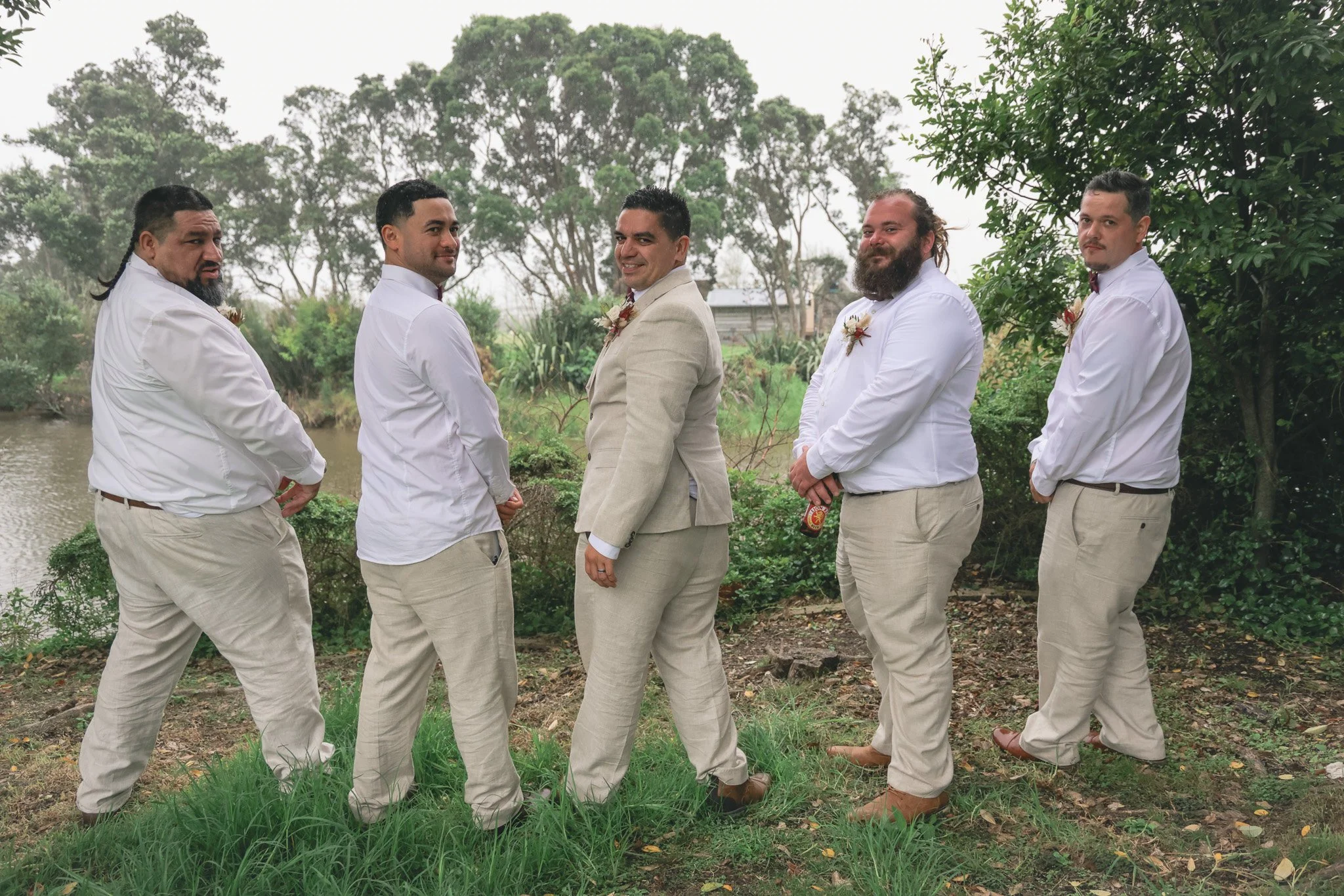 Six men dressed in suits and white shirts standing outdoors near a river with trees in the background.