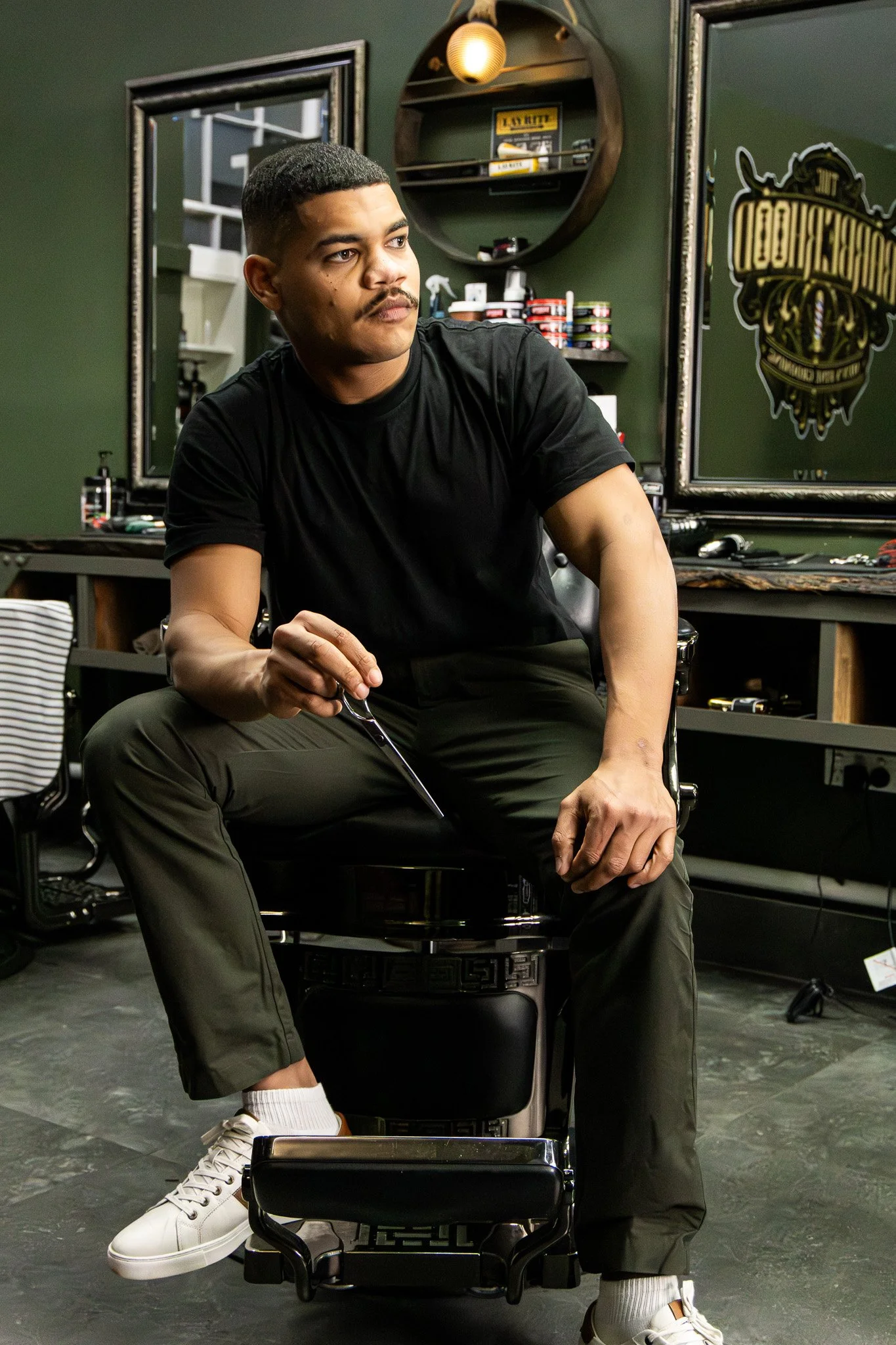 A man sitting on a black chair with one leg crossed over the other, holding glass in one hand. He has short hair, a mustache, and is wearing a black T-shirt, dark pants, white socks, and white sneakers. The background includes mirrors, shelves with h