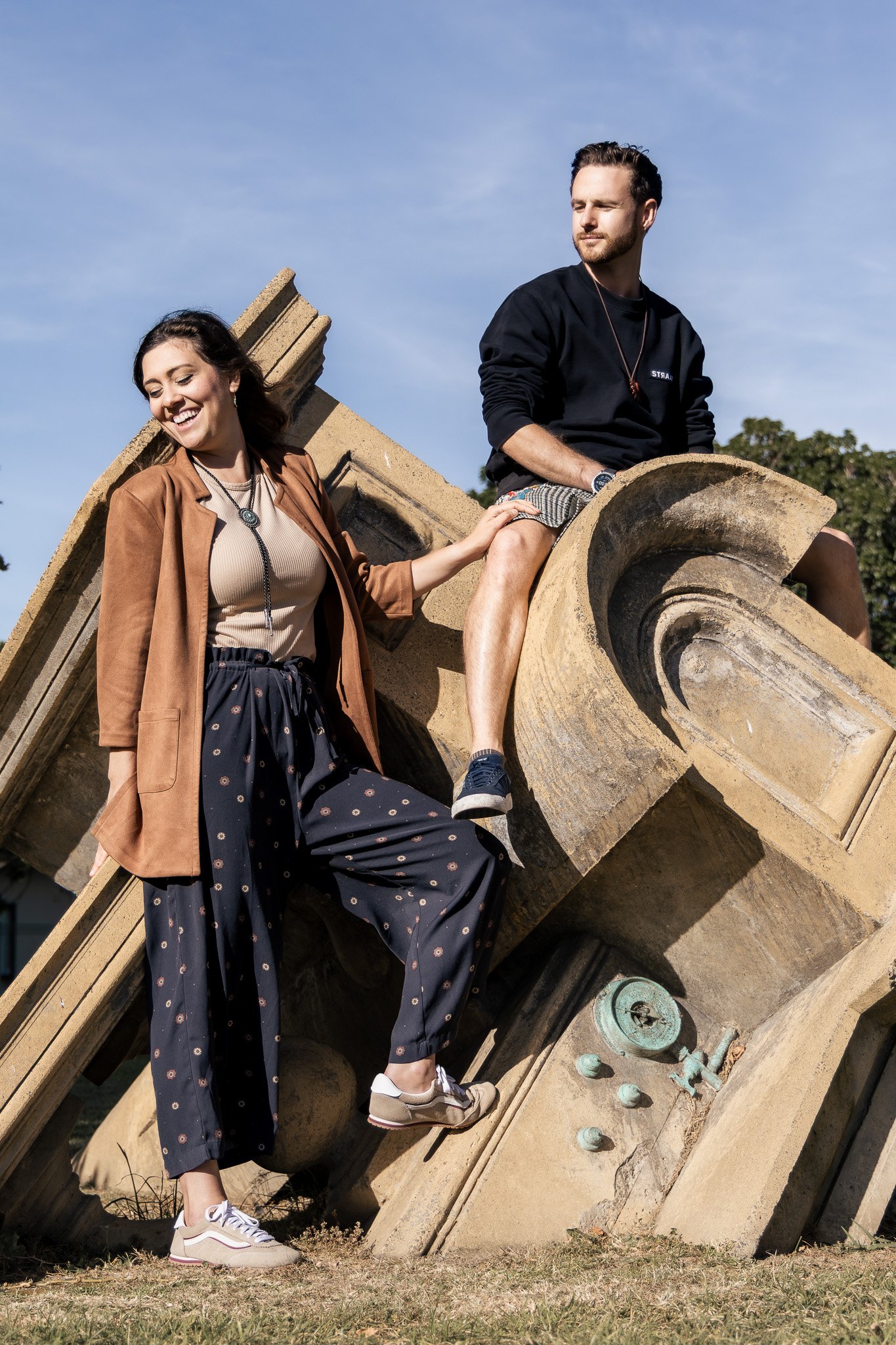 A man sitting on a decorative stone structure outdoors, with a woman touching his knee. The woman smiles while the man looks into the distance. They are dressed casually and the scene has a bright, clear sky.