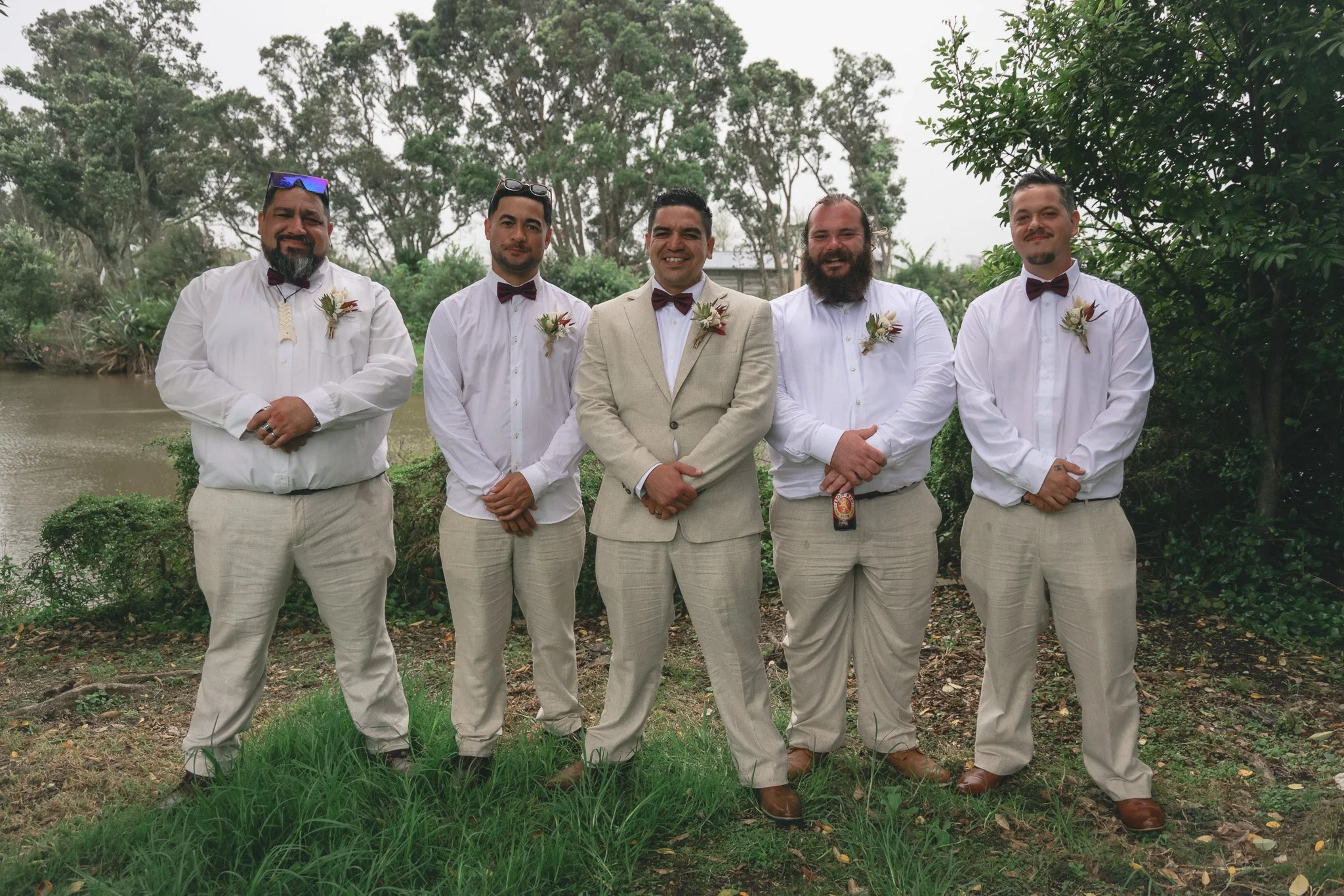 A group of five men dressed in formal attire, wearing white shirts, beige pants, brown bow ties, and boutonnières, standing outdoors with trees and a body of water in the background, smiling for a photo.