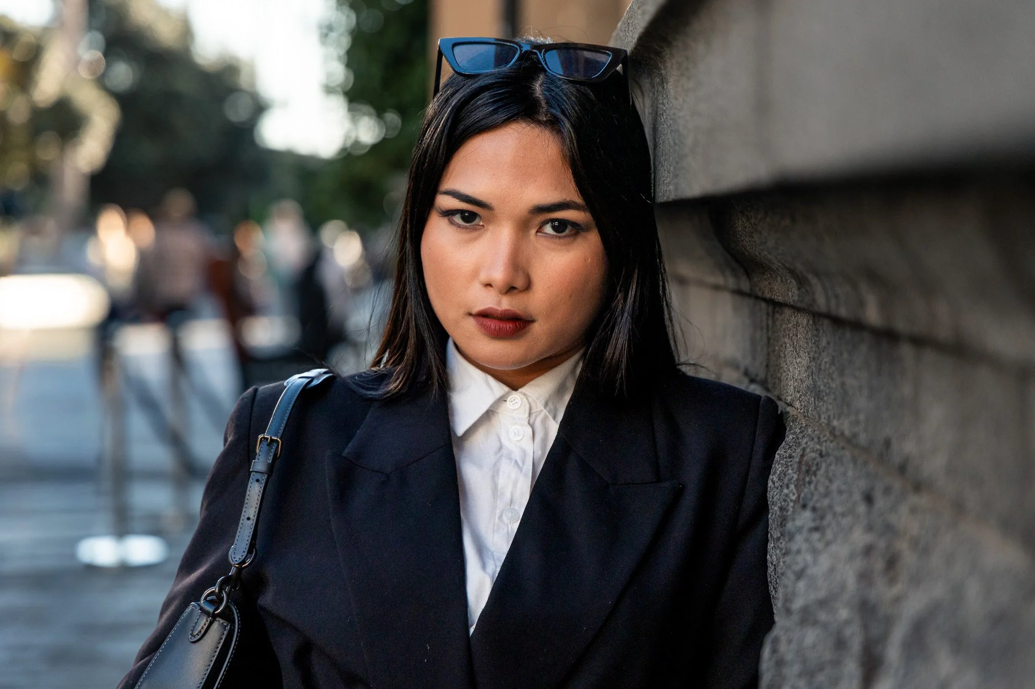 A woman with long dark hair wearing a white collared shirt, dark blazer, and glasses on her head, standing against a stone wall on a city street.