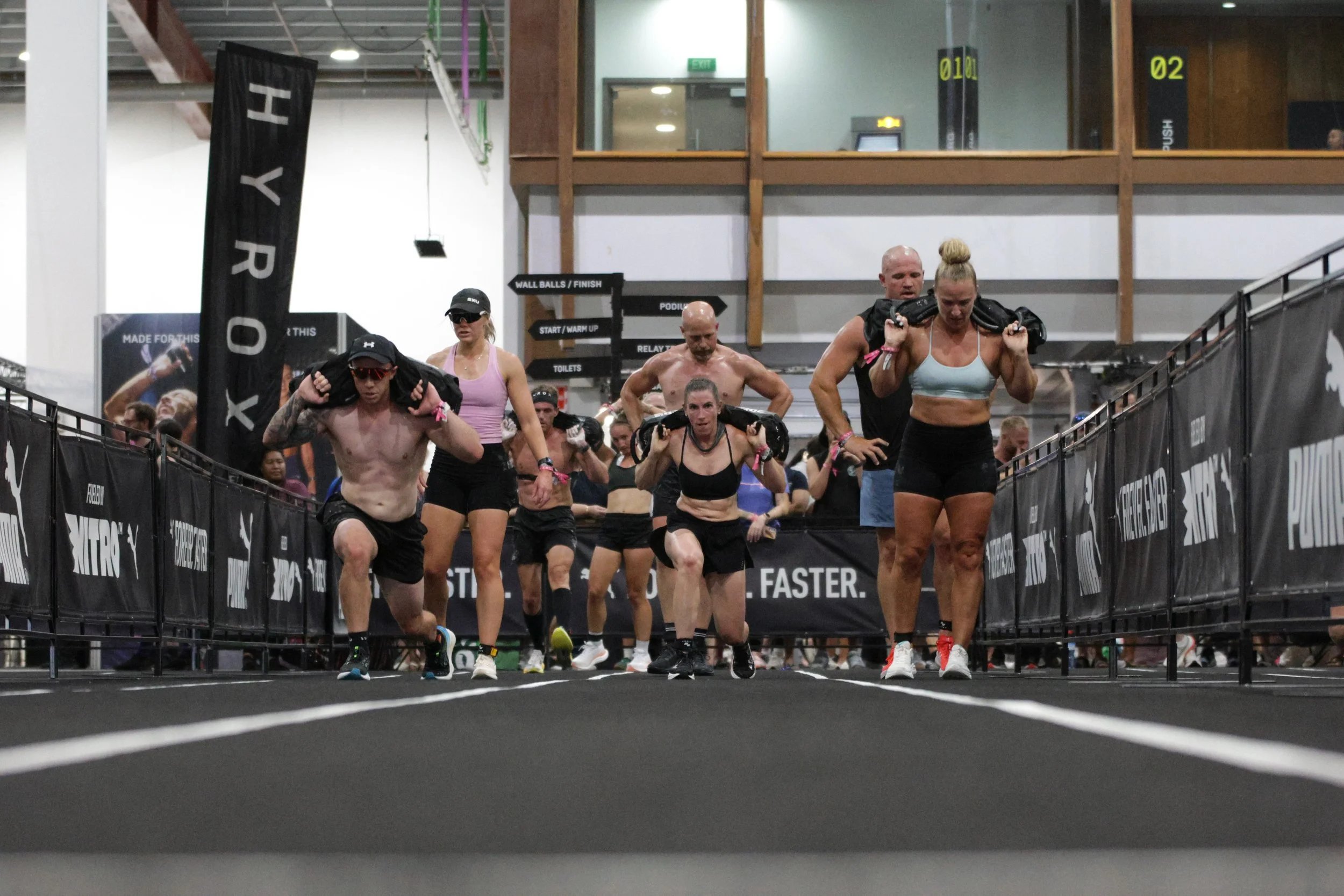 Group of people participating in a fitness event at an indoor gym, carrying sandbags on their shoulders and running on a treadmill-like surface, with black barriers on the sides.