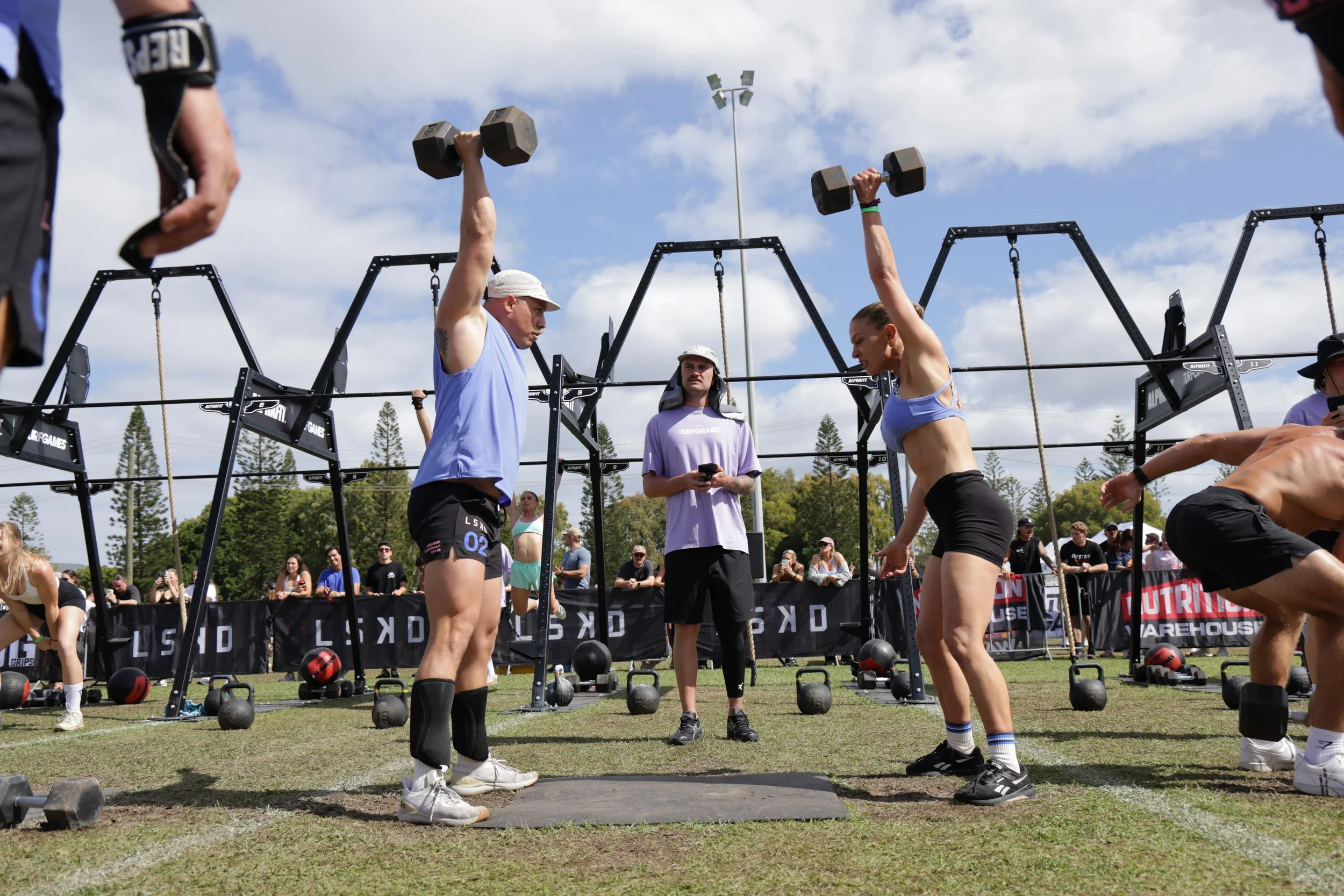 People participating in an outdoor fitness competition, lifting dumbbells overhead, with spectators watching in the background, and workout equipment on the ground. The setting is a grassy field under a partly cloudy sky.