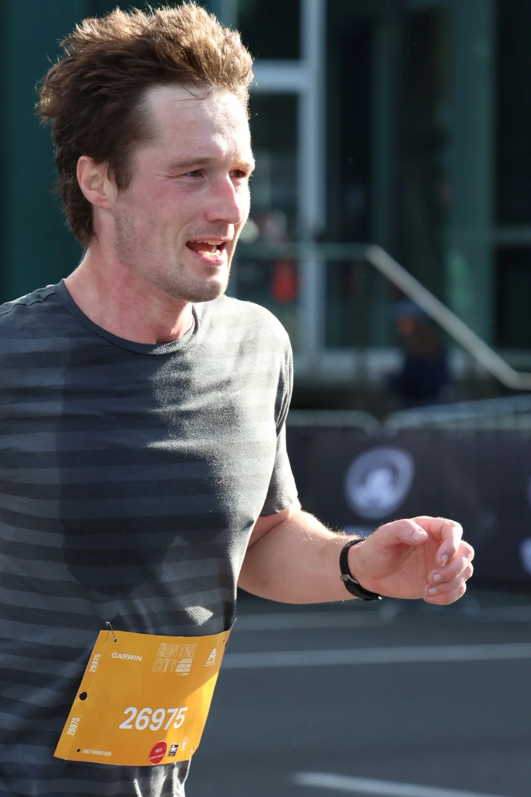 A man running in a race, wearing a gray striped shirt and a yellow bib with the number 26975, appears to be tired or exhausted, with a focused expression on his face.