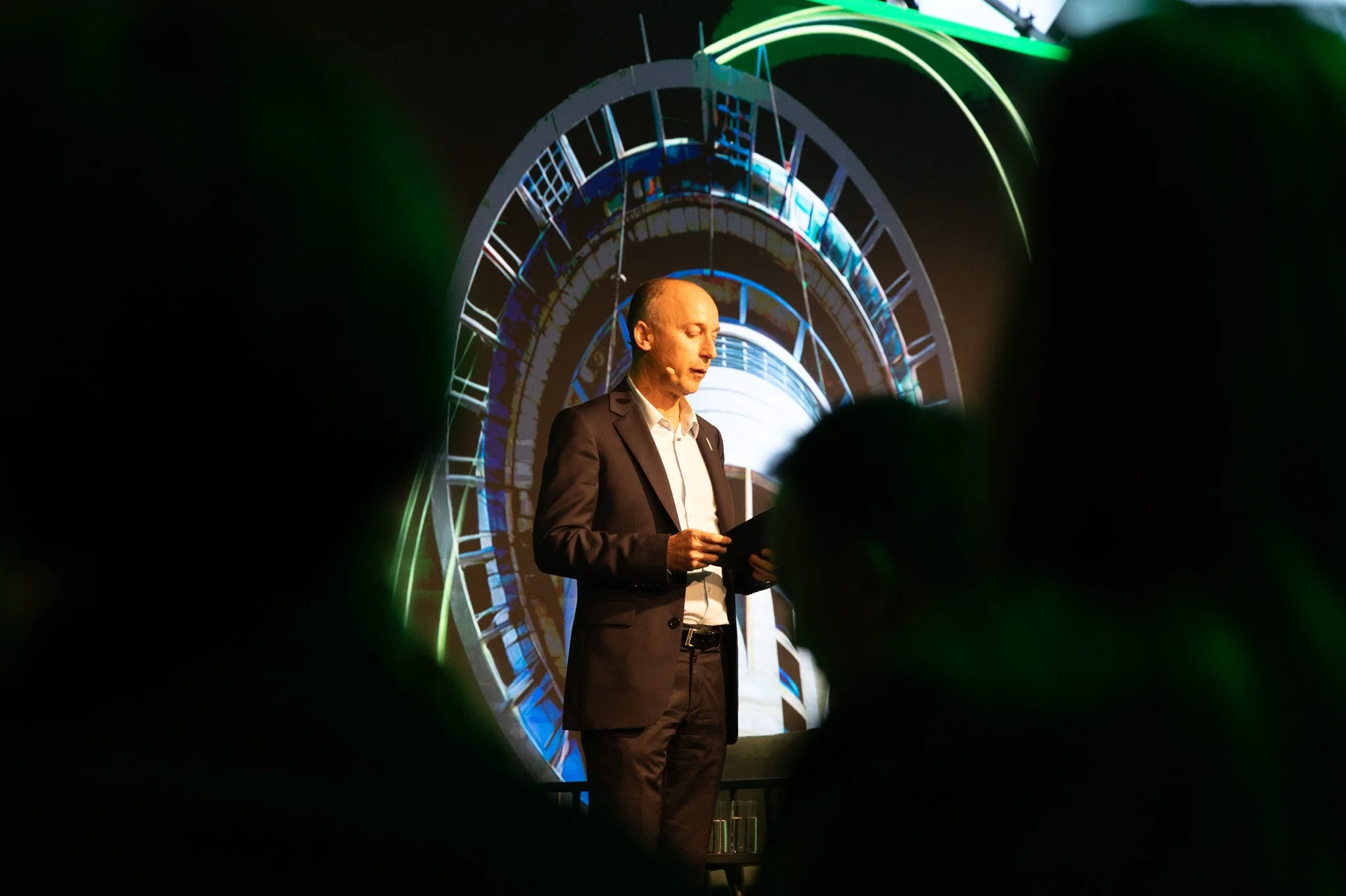 A man in a suit is standing on a stage, reading from a tablet, with a colorful circular digital display behind him. There are silhouettes of audience members in the foreground.