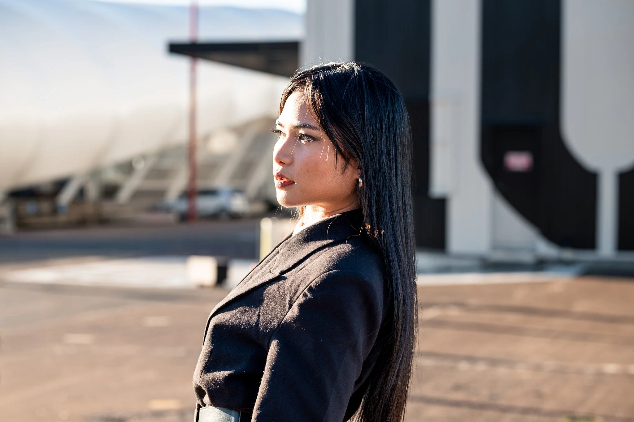 A woman with long black hair and earrings looking to the side outdoors during daytime, with a blurred background of buildings and a parking lot.