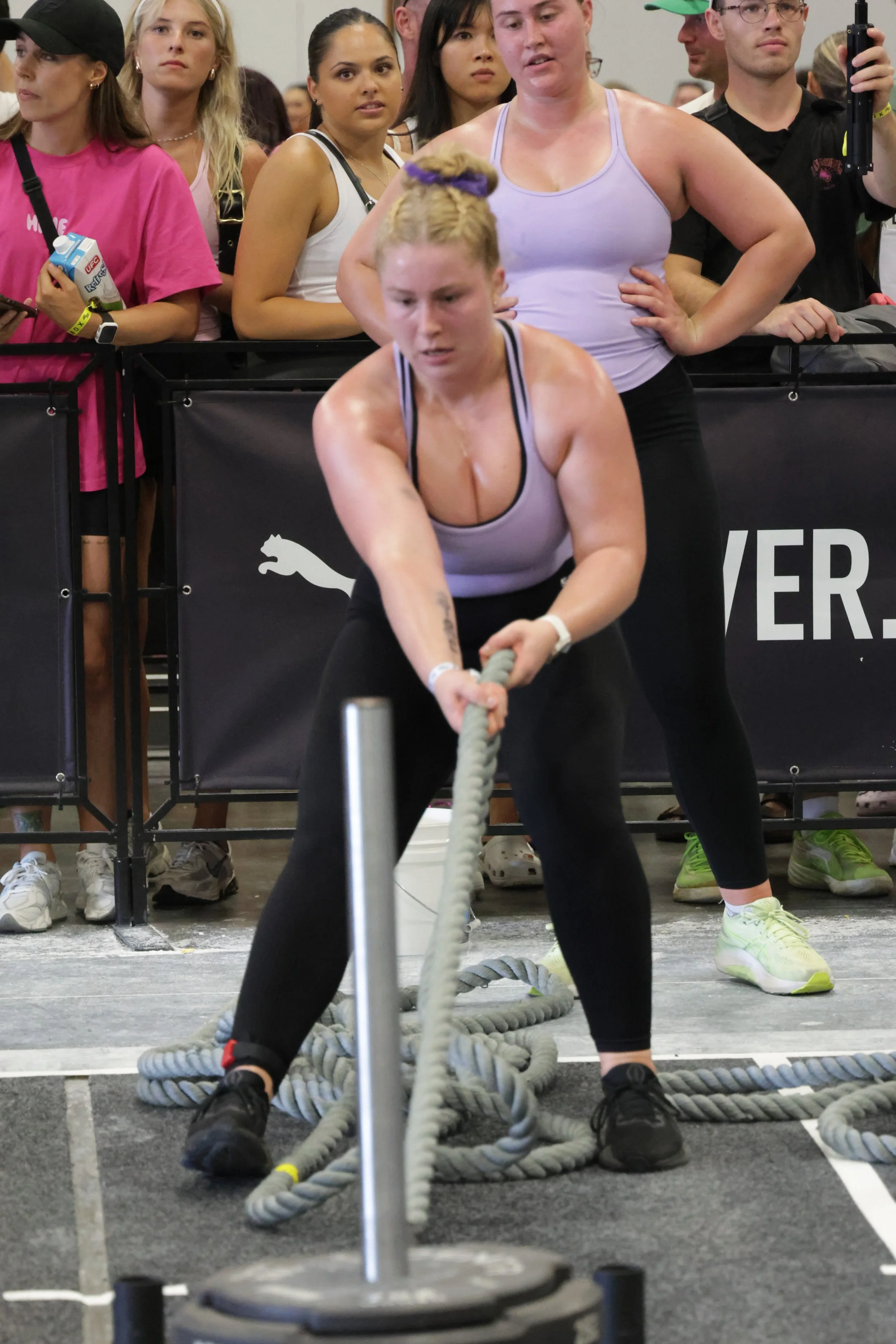 A woman in athletic gear pulling on thick battle ropes in a gym, with several spectators or participants behind her watching attentively.